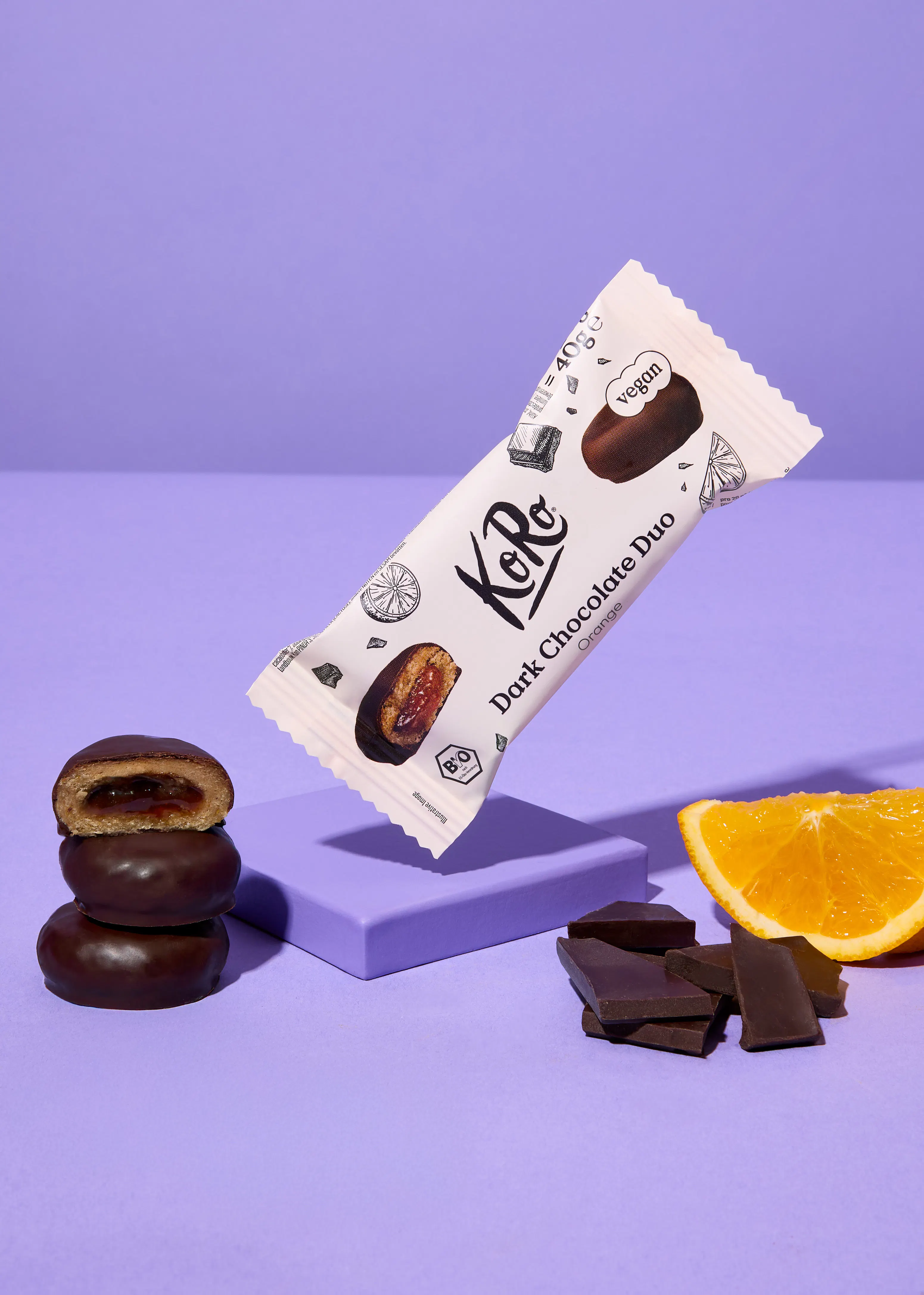eine koro dark chocolate duo orange riegelverpackung schwebt über einem stapel schokoladenkeksen mit orangenfüllung, einer orangenscheibe und schokoladenstücken auf einem lila hintergrund.