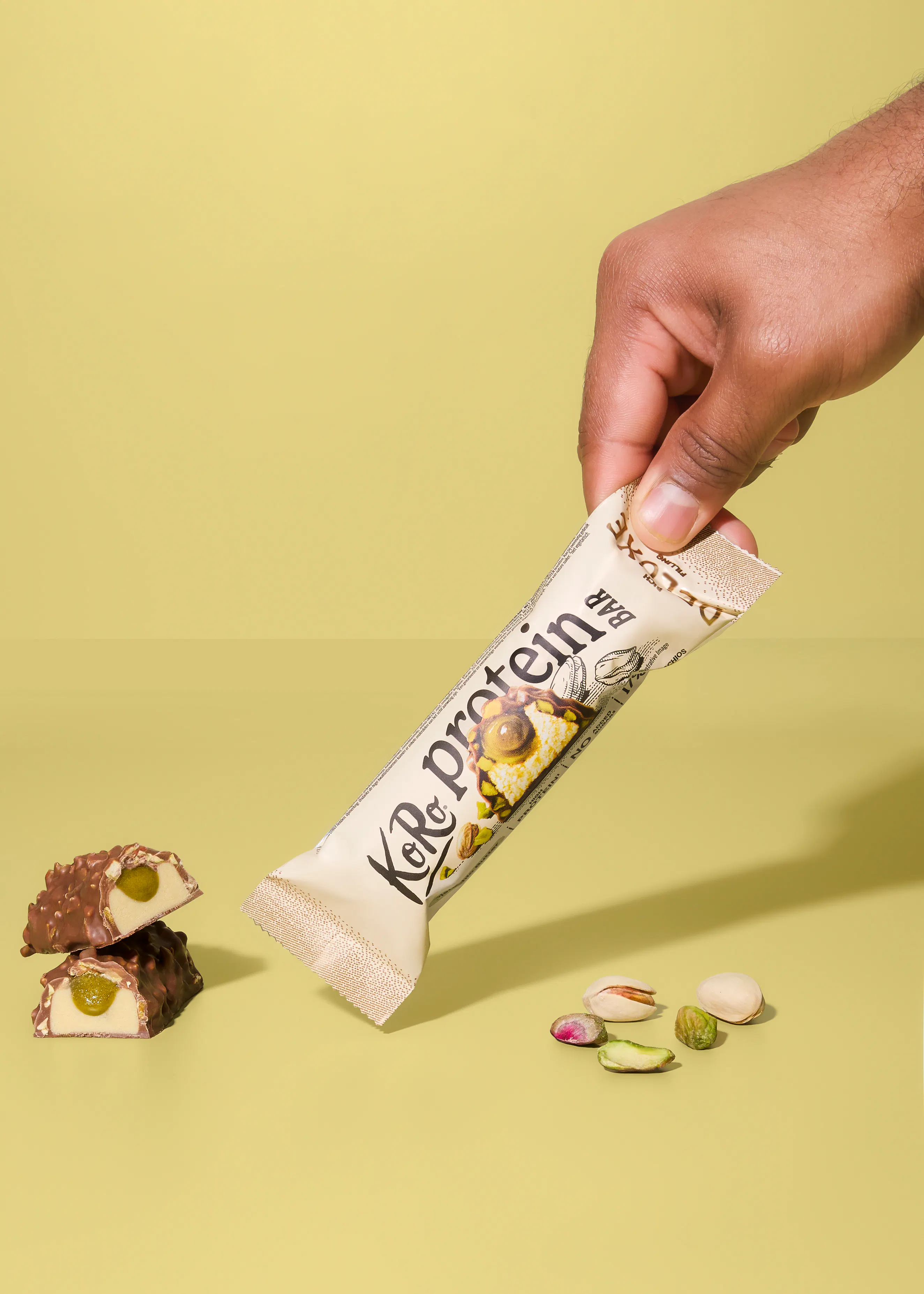 una mano tiene una barretta proteica ko&protein, con due pezzi tagliati che mostrano un ripieno al pistacchio e alcuni pistacchi sparsi su uno sfondo verde chiaro.
