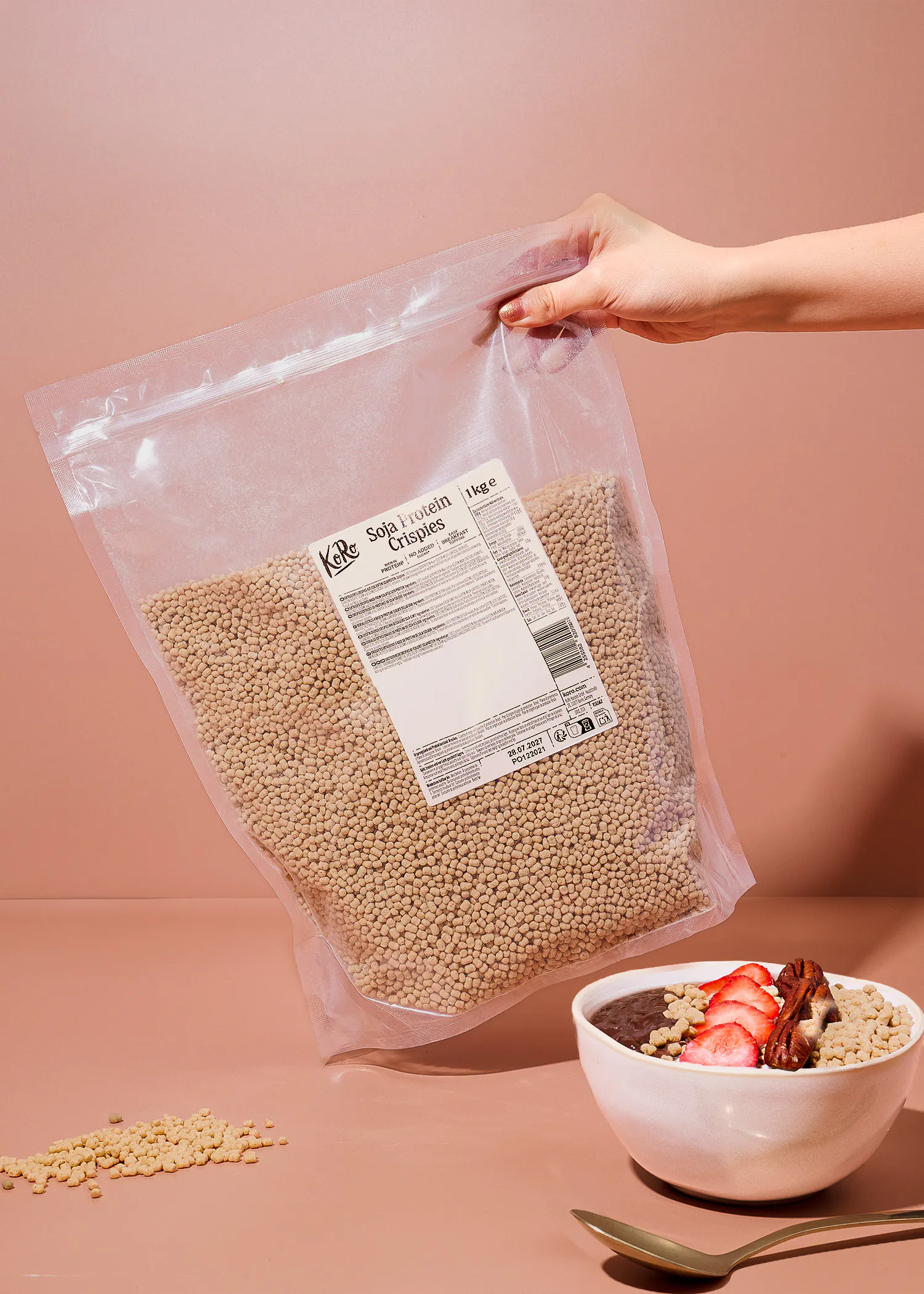 una mano tiene una confezione da un chilogrammo di soia protein crispies koro accanto a una ciotola di colazione con fragole e noci.