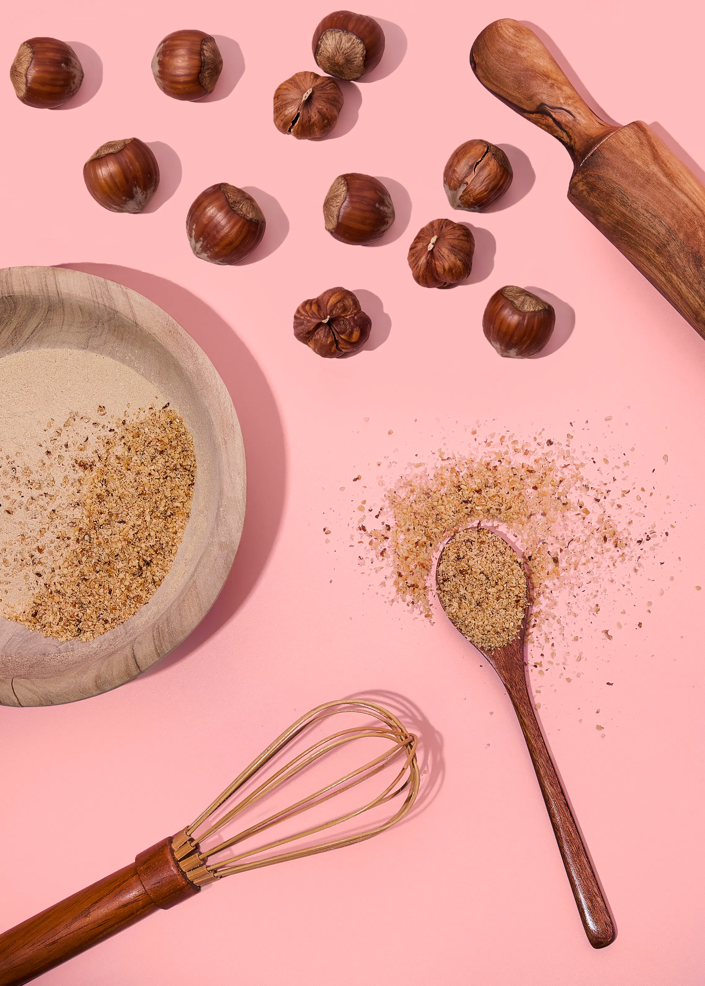 auf einem rosa hintergrund sind haselnüsse, gemahlene haselnüsse in einer holzschale und auf einem holzlöffel, ein nudelholz und ein schneebesen arrangiert.