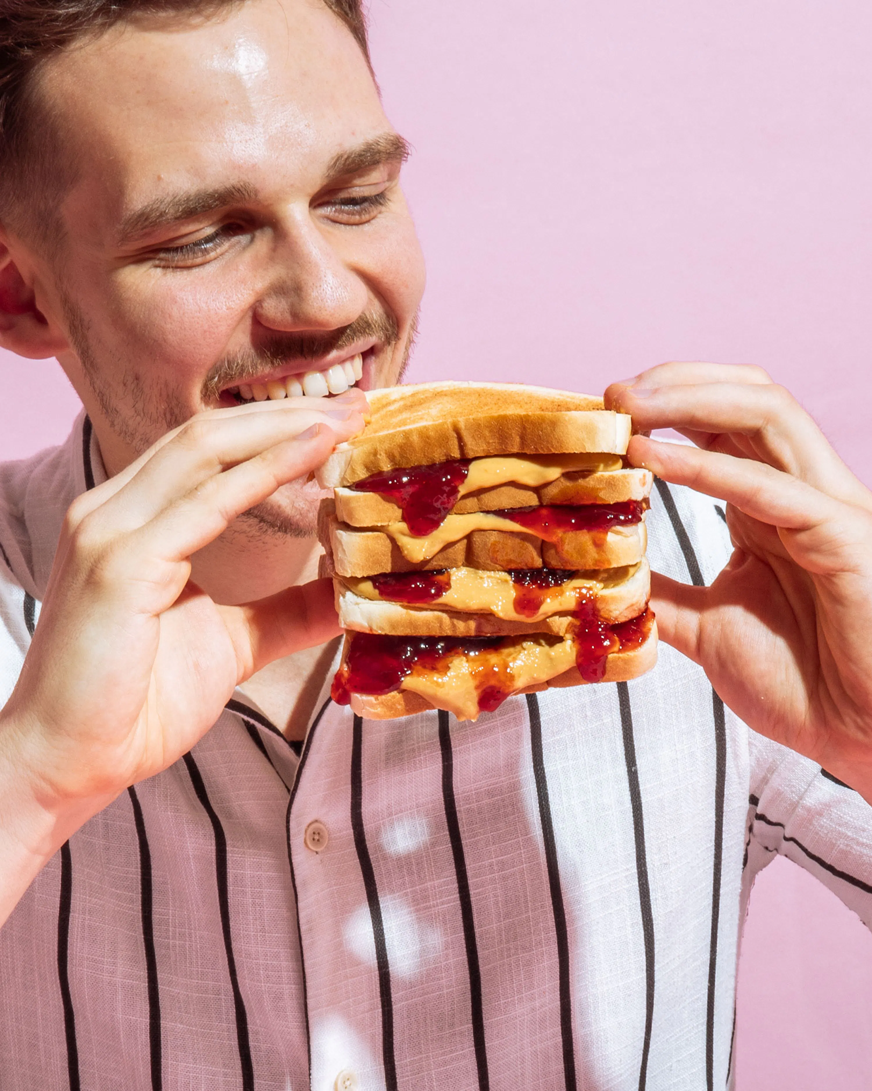 ein lächelnder mann mit bartstoppeln hält ein mehrschichtiges erdnussbutter- und marmeladenbrot und ist bereit, hineinzubeißen.