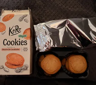 una confezione di biscotti vegani koro al burro di arachidi accanto a un vassoio di plastica contenente biscotti rotondi.