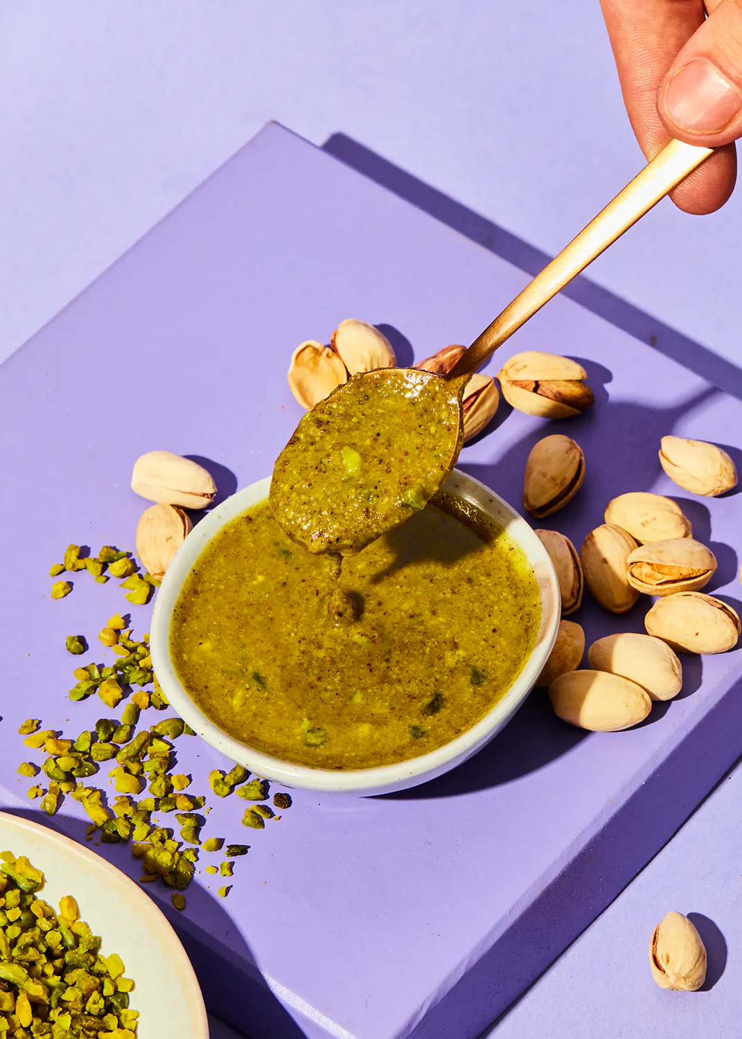 une cuillère remplie de sauce verte est sur une table violette entourée de pistaches
