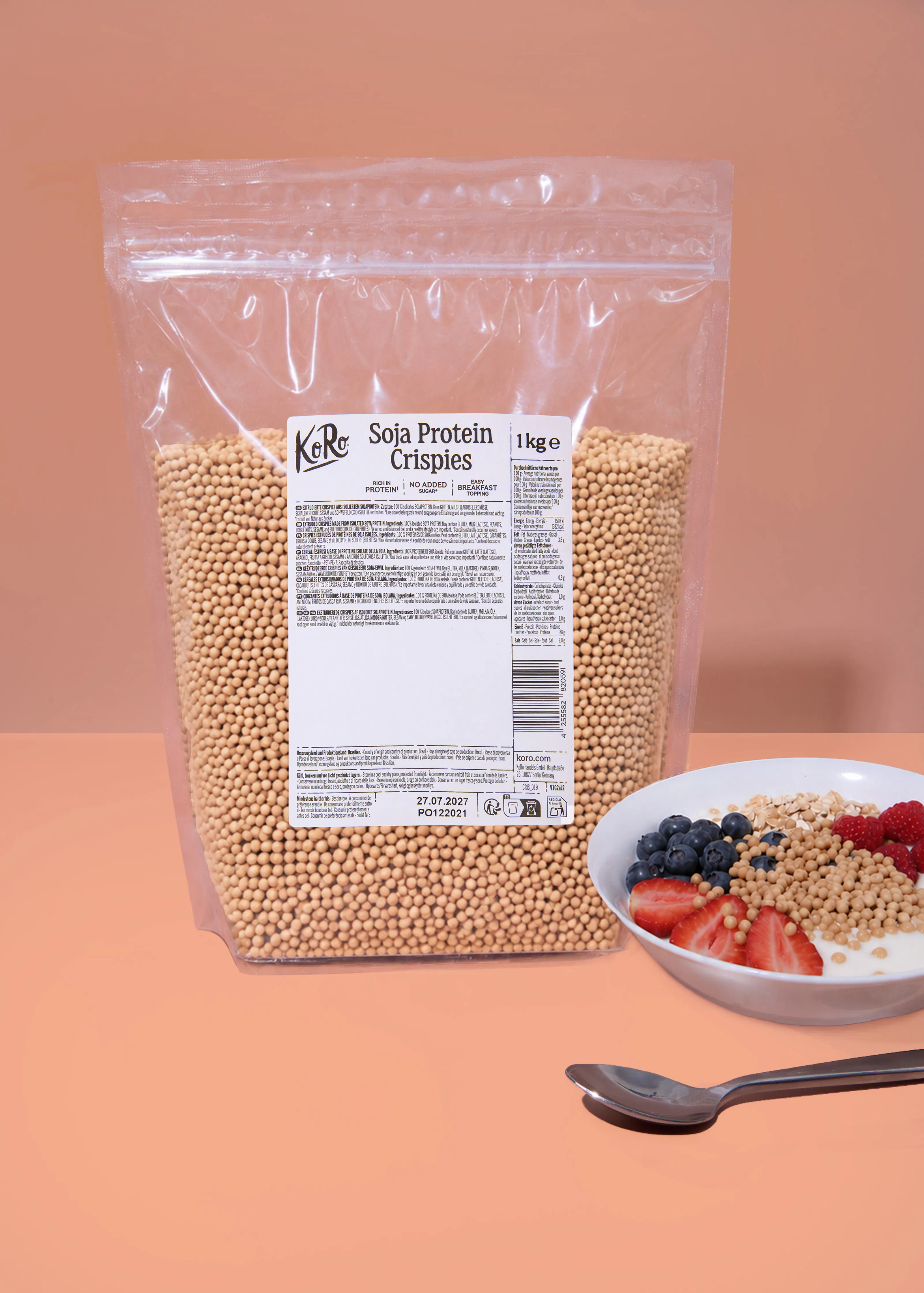 eine große, durchsichtige tüte koro soja protein crispies steht neben einer schale joghurt mit crispies, erdbeeren, heidelbeeren und haferflocken.