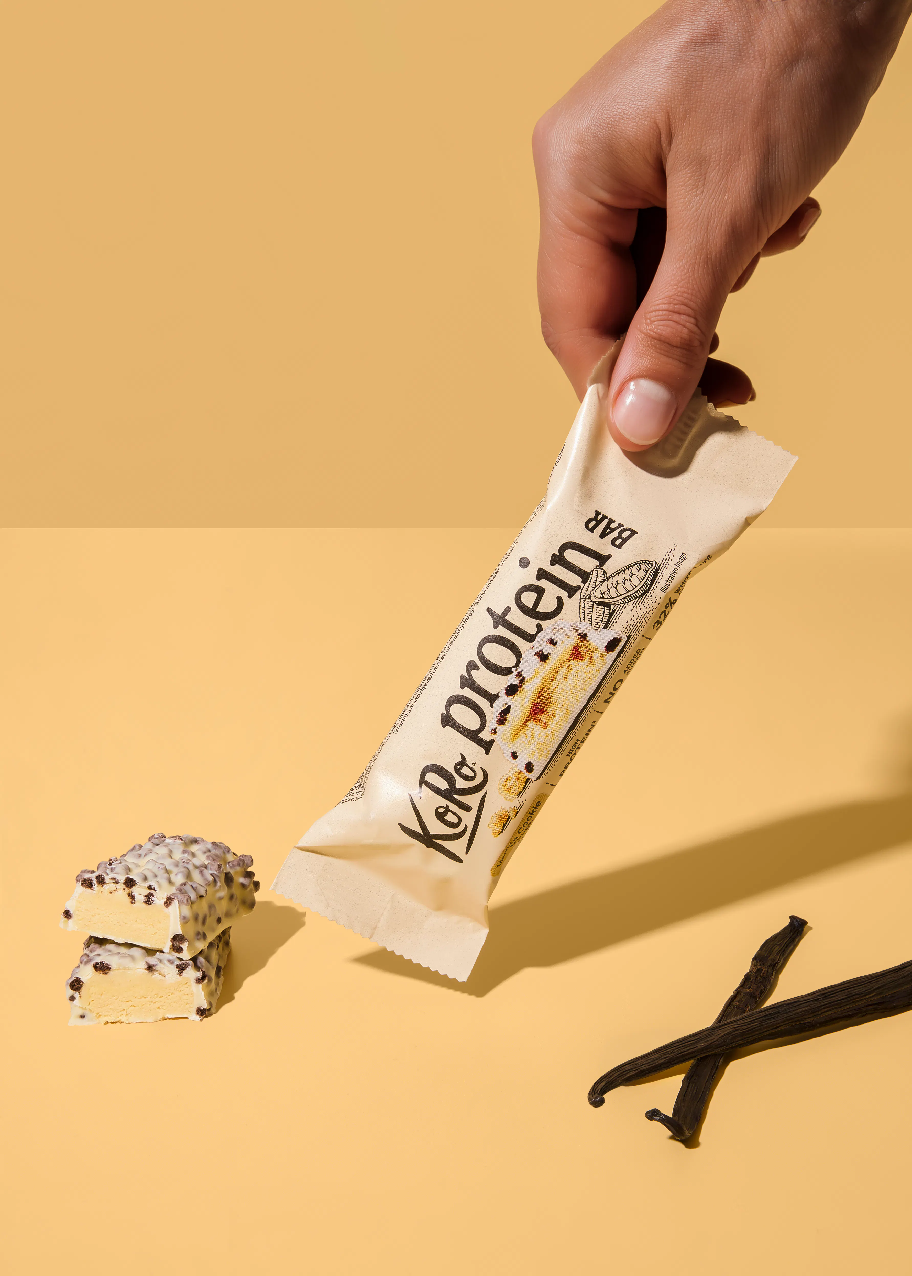 una mano tiene una barretta proteica koro al gusto biscotto alla vaniglia, con due pezzi tagliati della barretta e due baccelli di vaniglia su uno sfondo giallo.