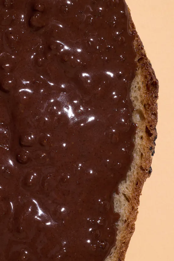 une tranche de pain recouverte d'une pâte à tartiner au chocolat brillante, avec la croûte du pain visible sur le côté droit.