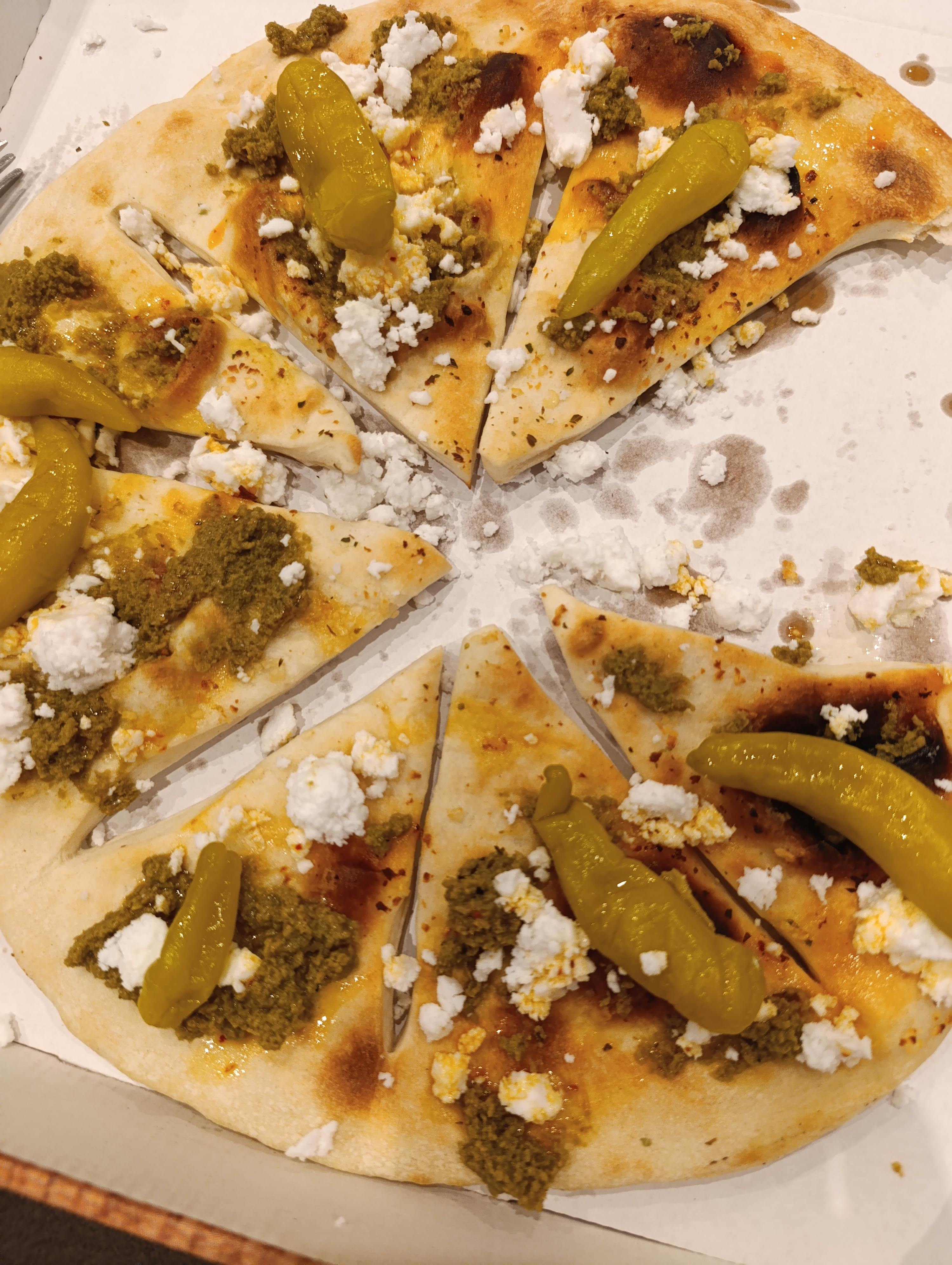 eine teilweise gegessene pizza mit pesto, zerbröseltem käse und grünen peperoni, in scheiben geschnitten in einer pizzabox.