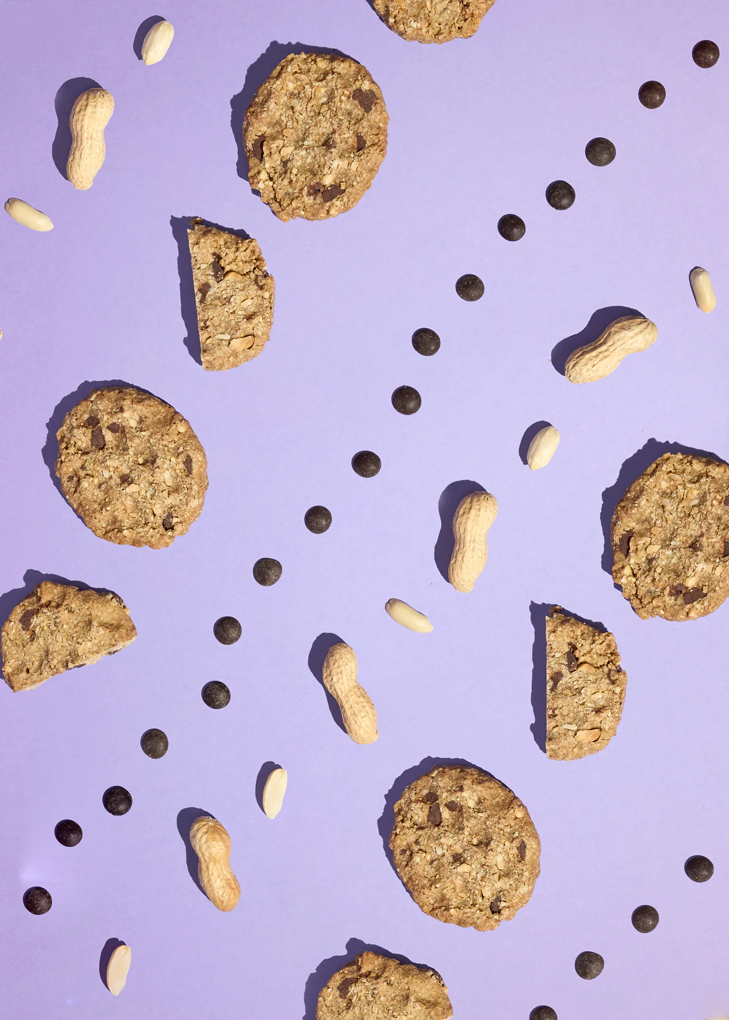 des biscuits aux flocons d'avoine et pépites de chocolat, des cacahuètes entières et décortiquées, et des pépites de chocolat sont disposés de manière éparpillée sur un fond violet.