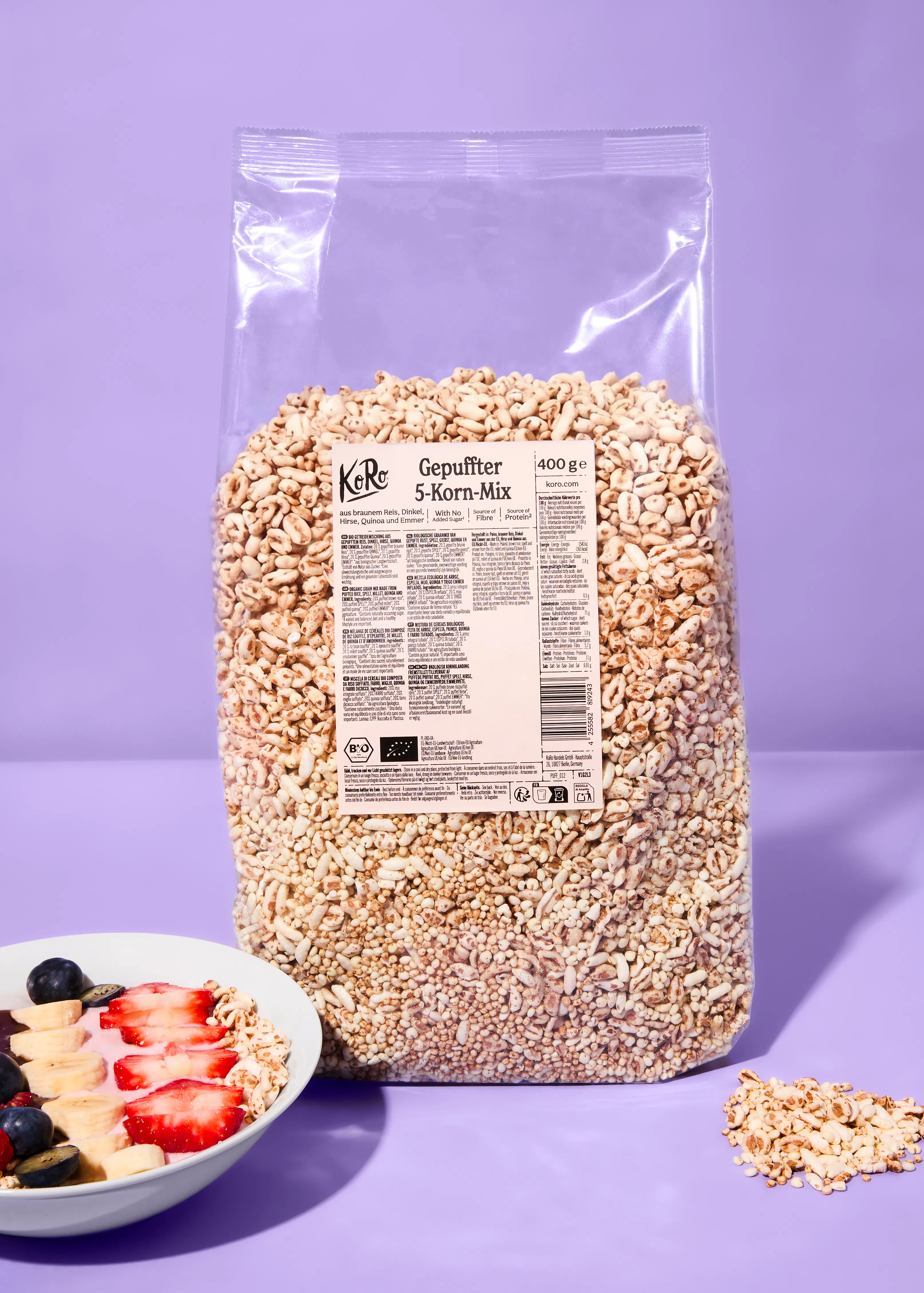 a large clear bag of koro gepuffter 5-korn-mix (puffed 5-grain mix) stands on a purple surface next to a bowl of yogurt topped with fruit and some spilled cereal.