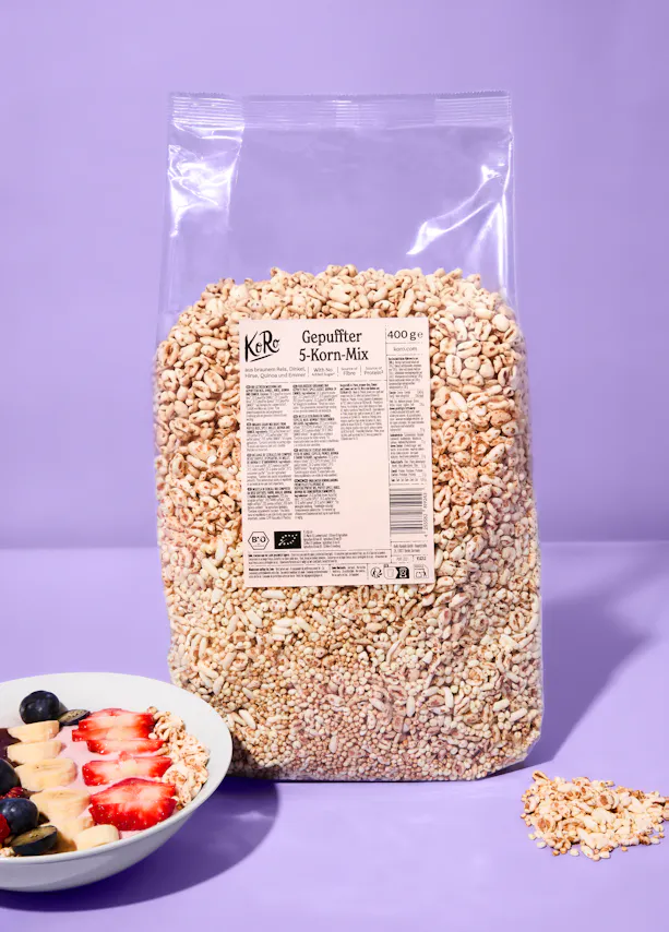 a large clear bag of koro gepuffter 5-korn-mix (puffed 5-grain mix) stands on a purple surface next to a bowl of yogurt topped with fruit and some spilled cereal.