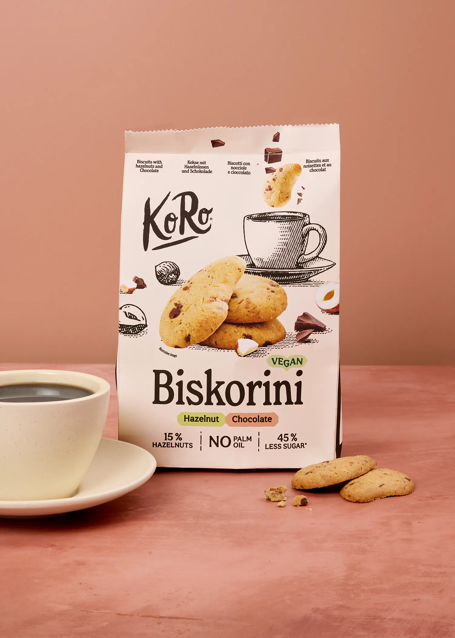 eine weiße verpackung von koro biskorini veganen haselnuss-schokoladenkeksen steht neben einer tasse kaffee und einigen keksen auf einer rosa oberfläche.