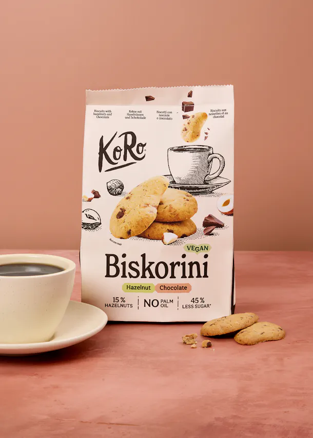 eine weiße verpackung von koro biskorini veganen haselnuss-schokoladenkeksen steht neben einer tasse kaffee und einigen keksen auf einer rosa oberfläche.