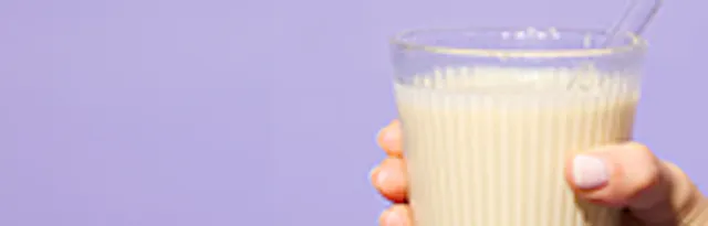 une main tient un verre de boisson pâle avec une paille devant un fond violet.
