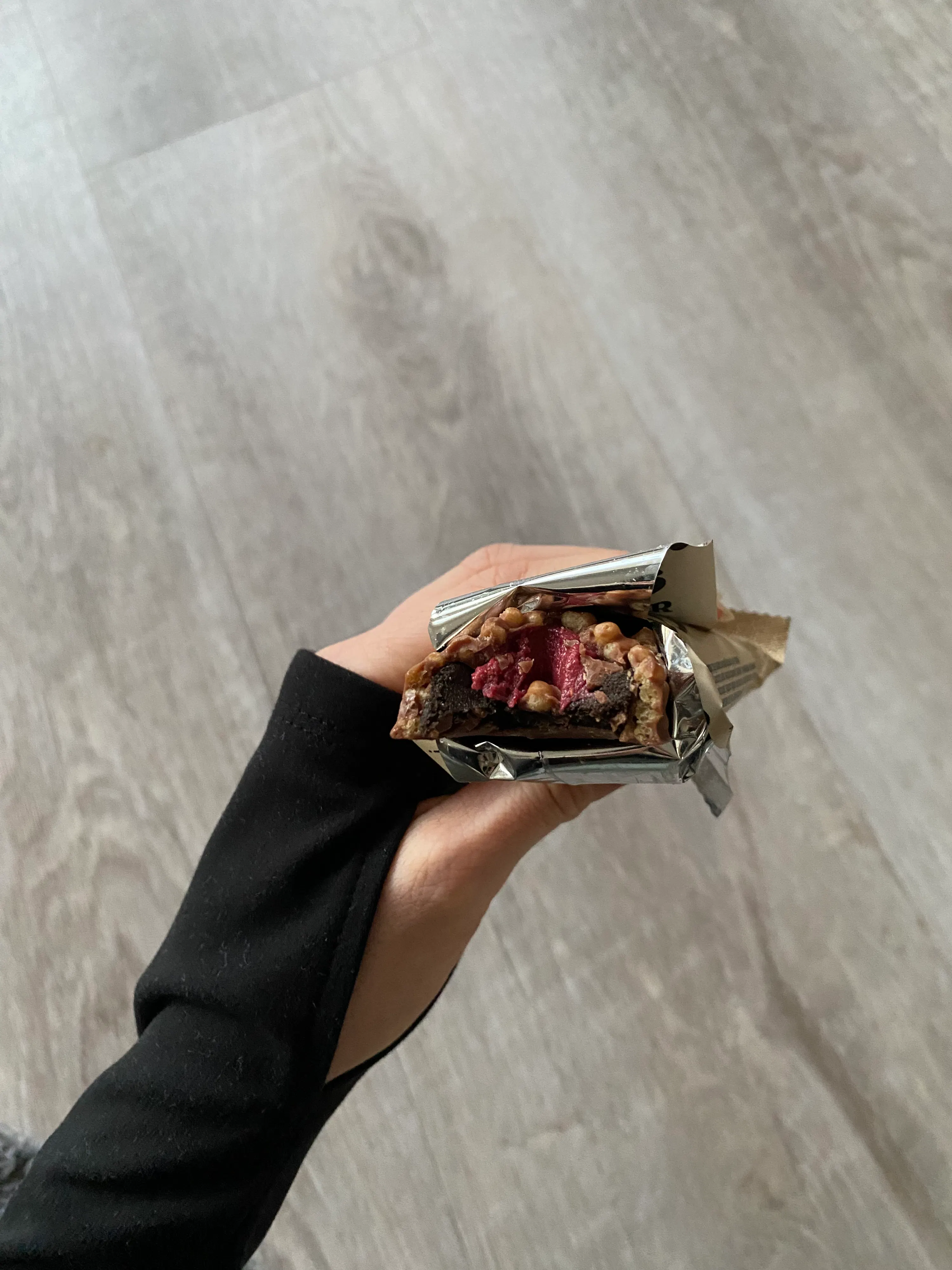 eine hand hält einen teilweise ausgepackten snackriegel mit dunkler basis, roten früchten und nüssen in silberner folie.