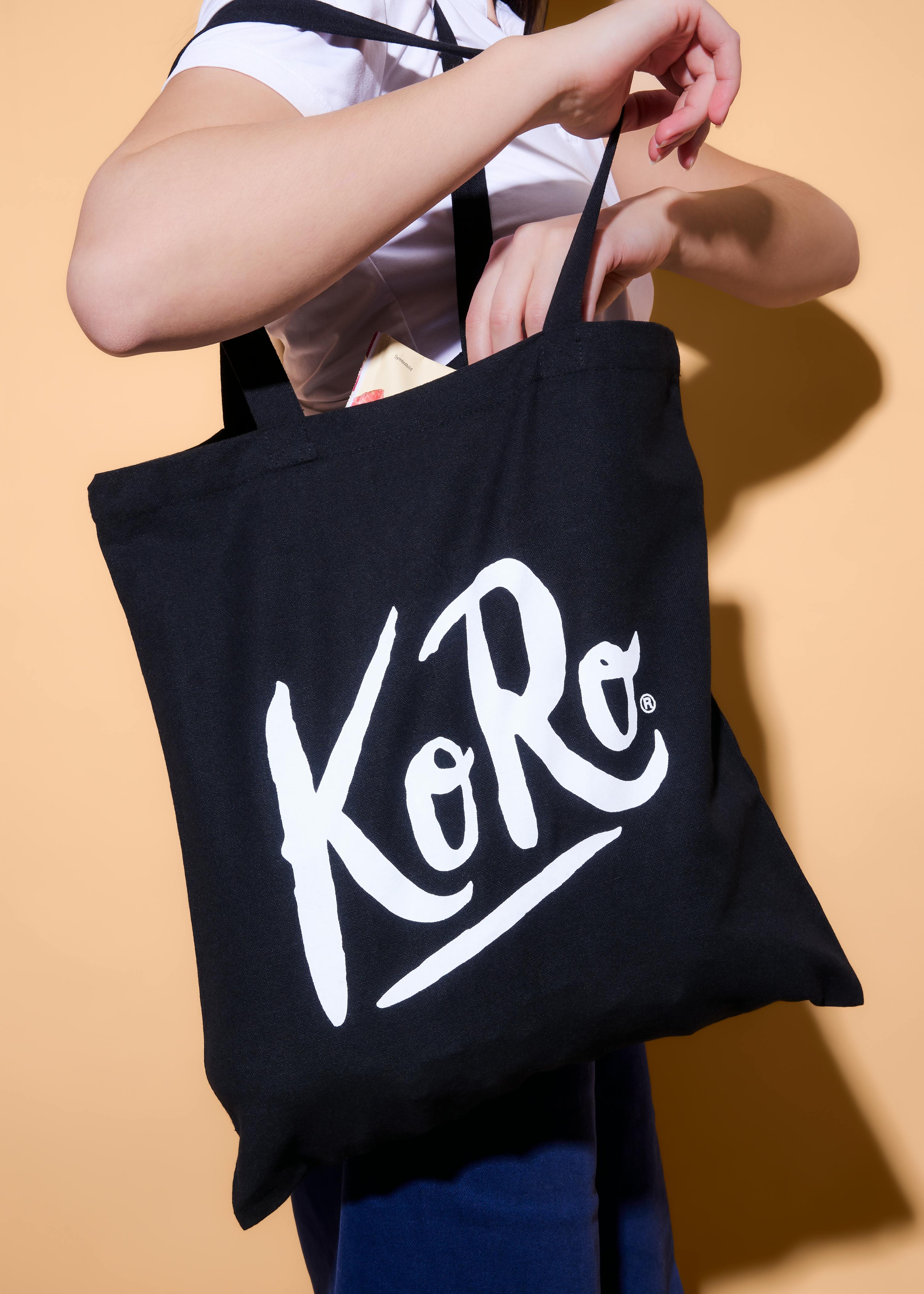 una persona tiene una borsa tote nera con il logo bianco 'koro' mentre mette o toglie qualcosa.