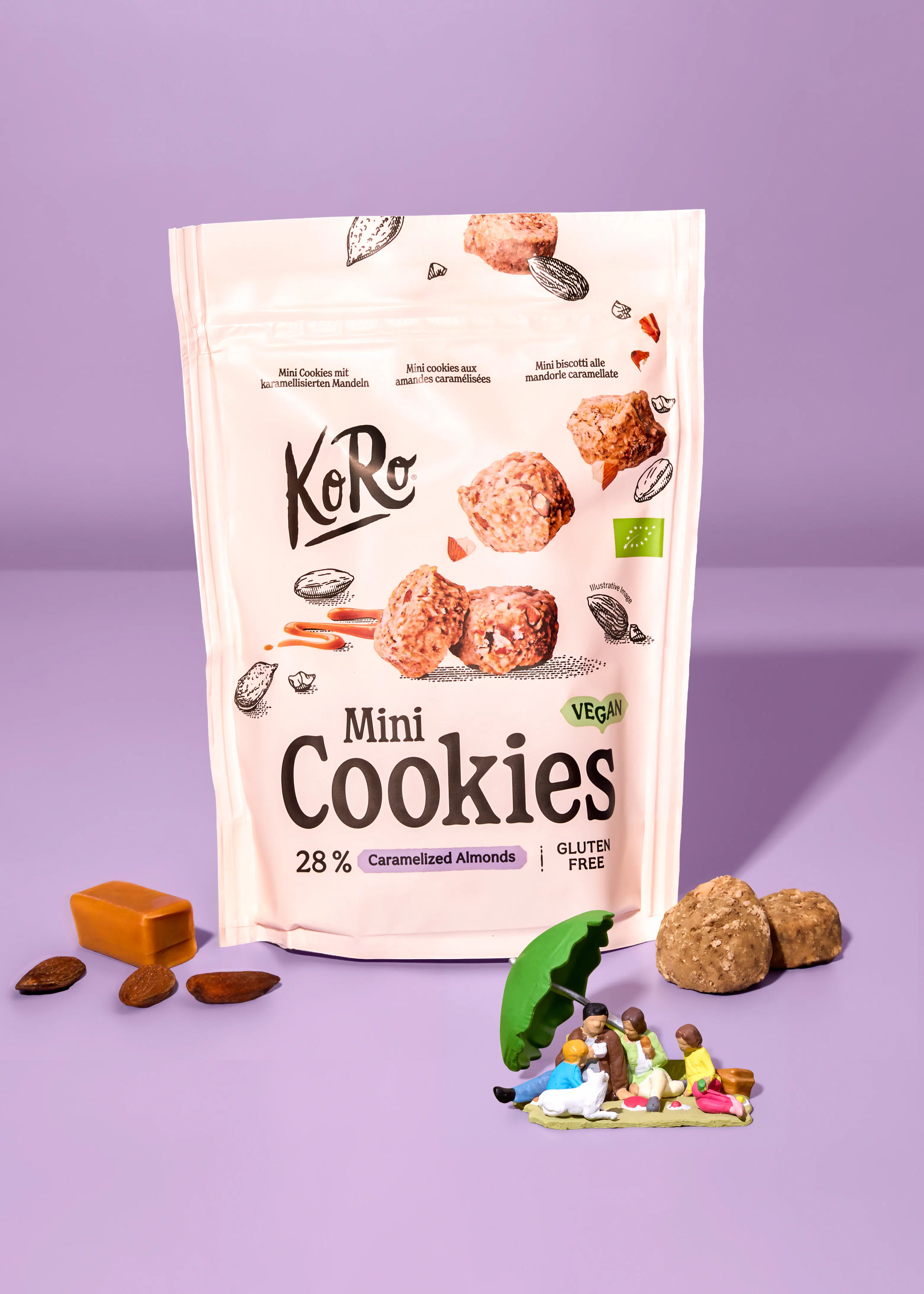 eine weiße verpackung von koro mini cookies mit karamellisierten mandeln, vegan und glutenfrei, umgeben von mandeln, karamell, kleinen keksen und einer miniatur-picknickszene auf lila hintergrund.