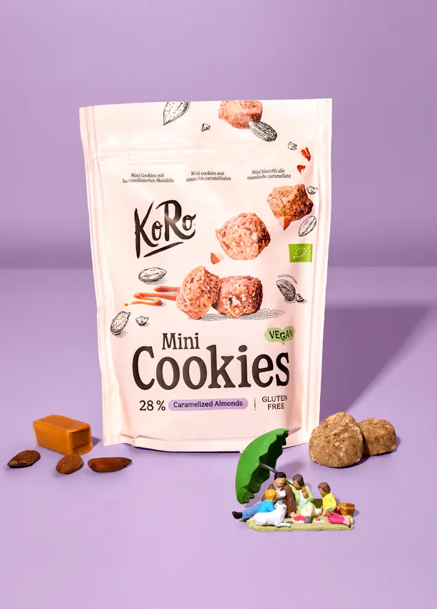 eine weiße verpackung von koro mini cookies mit karamellisierten mandeln, vegan und glutenfrei, umgeben von mandeln, karamell, kleinen keksen und einer miniatur-picknickszene auf lila hintergrund.