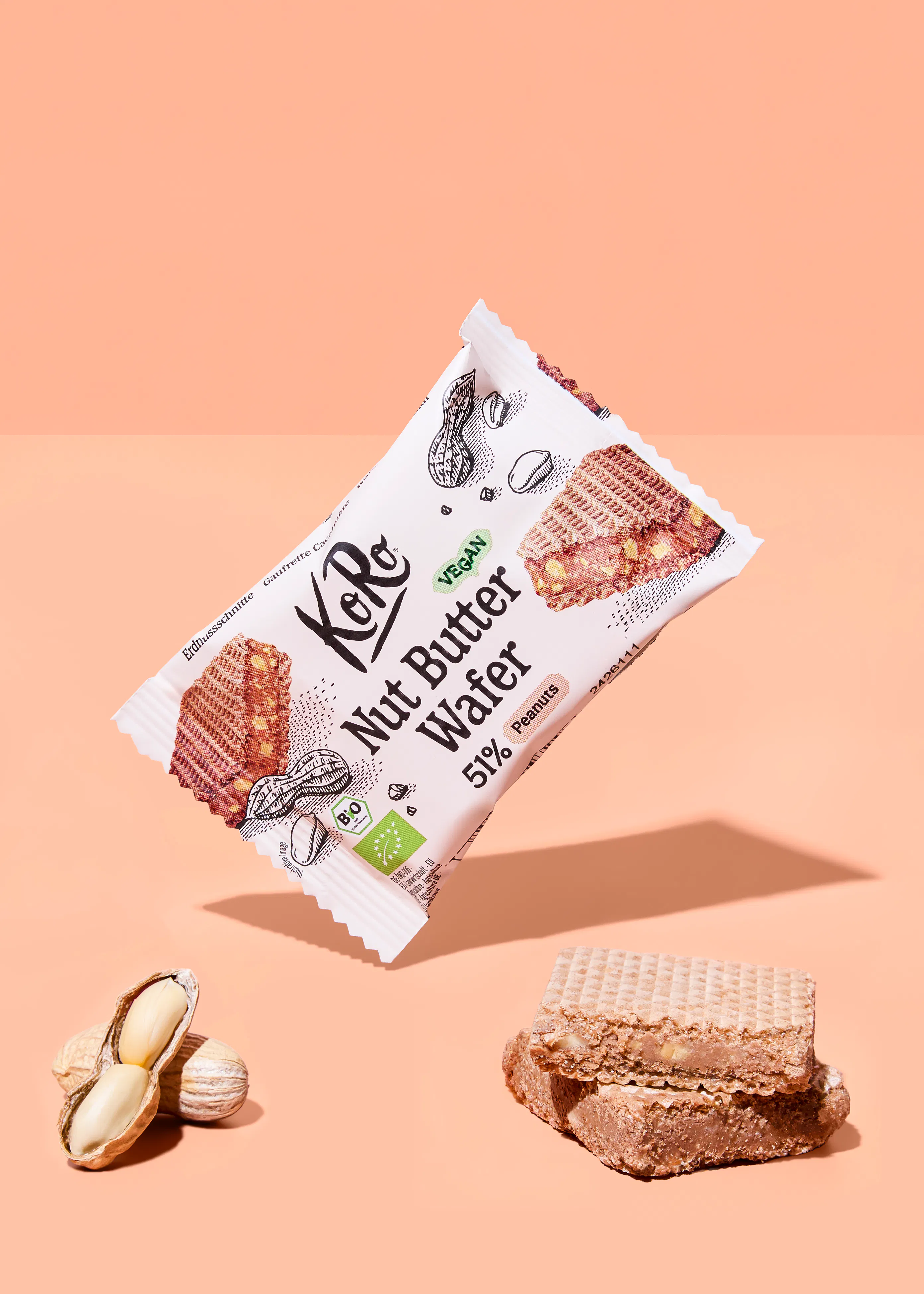 eine schwebende packung koro nut butter wafer, die als vegan und mit 51% erdnüssen gekennzeichnet ist, daneben eine offene erdnuss und zwei waffelstücke auf einem pfirsichfarbenen hintergrund.