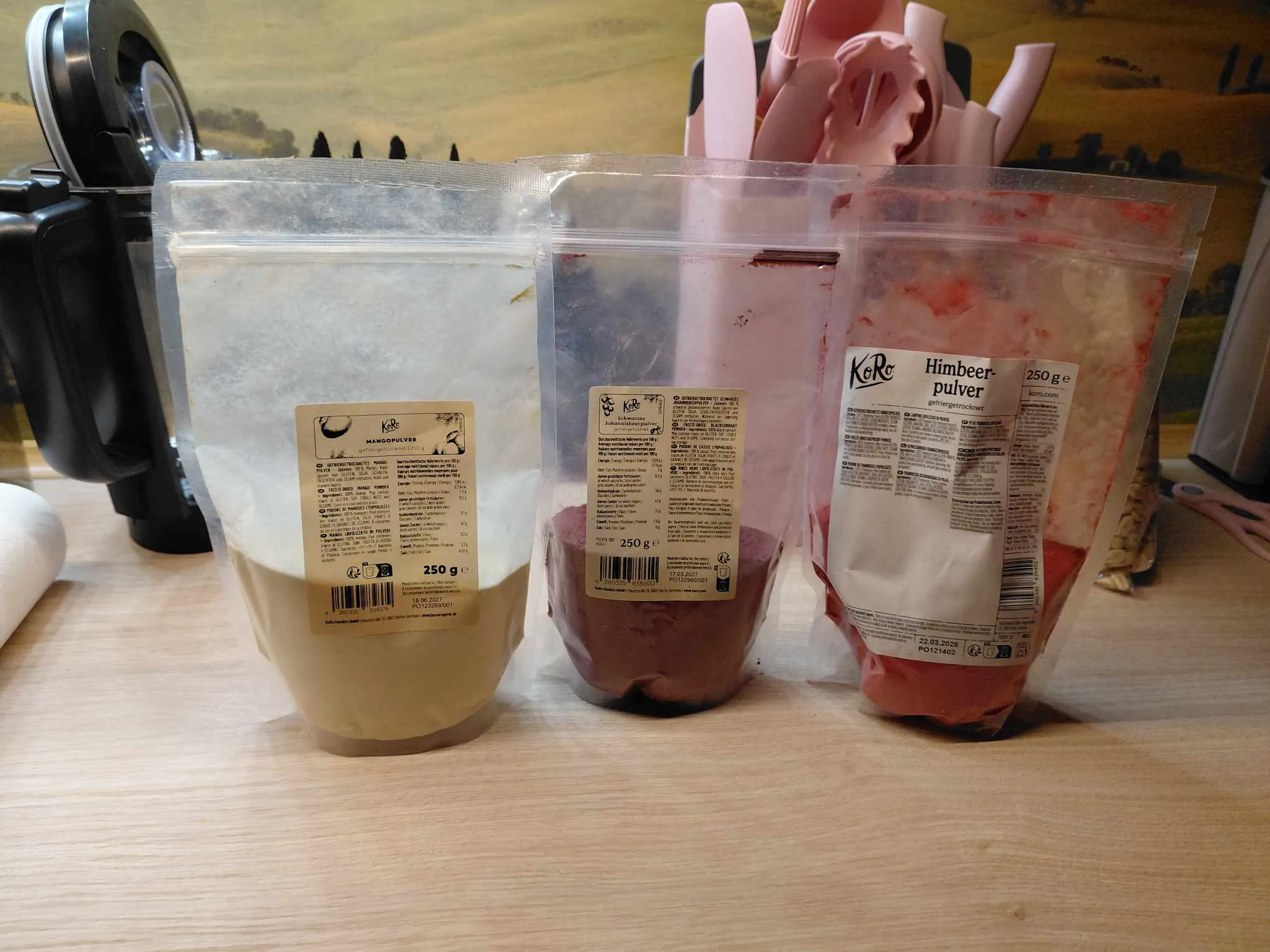 drei durchsichtige beutel mit koro-fruchtpulver in den sorten mango, schwarze johannisbeere und himbeere stehen nebeneinander auf einer küchenarbeitsplatte.