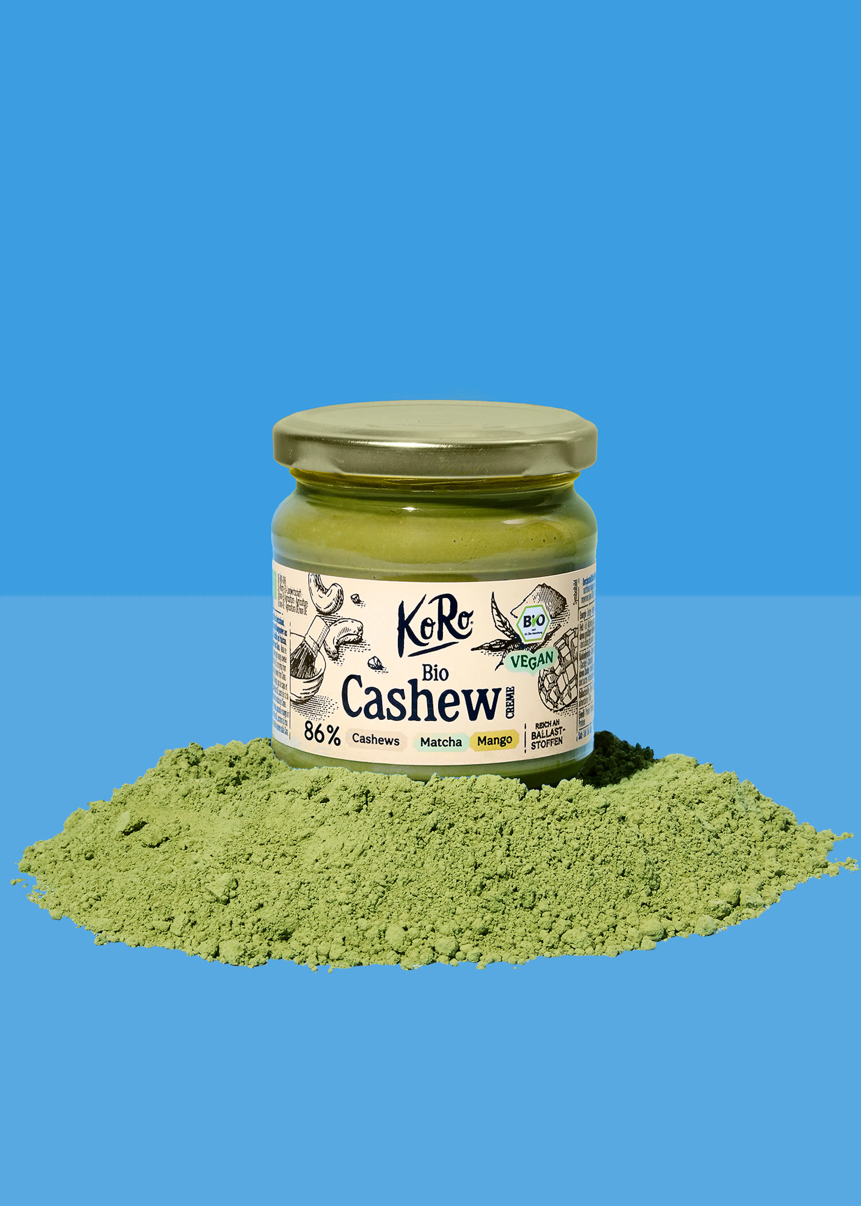 un barattolo di crema di anacardi bio koro al matcha e mango, con etichetta che indica "vegan" e "86% anacardi", è posizionato su una pila di polvere verde su uno sfondo blu.