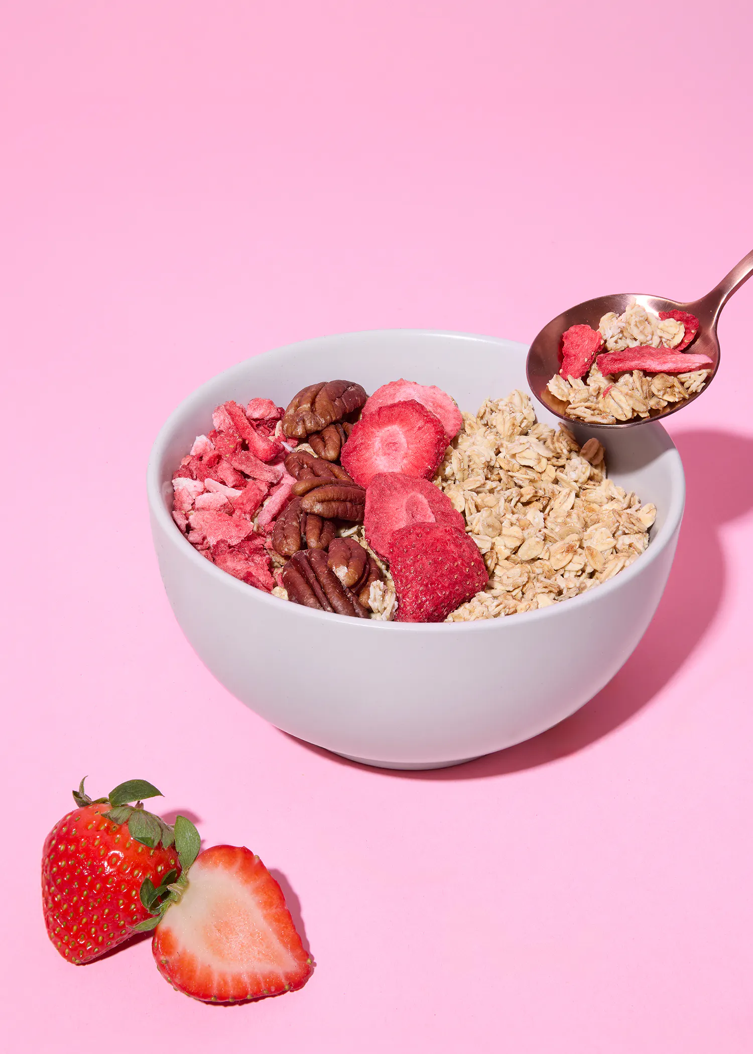 eine weiße schale mit haferflocken, pekannüssen und gefriergetrockneten erdbeeren steht auf einem pinkfarbenen hintergrund, während ein löffel mit haferflocken und erdbeeren über der schale schwebt und frische erdbeeren daneben liegen.