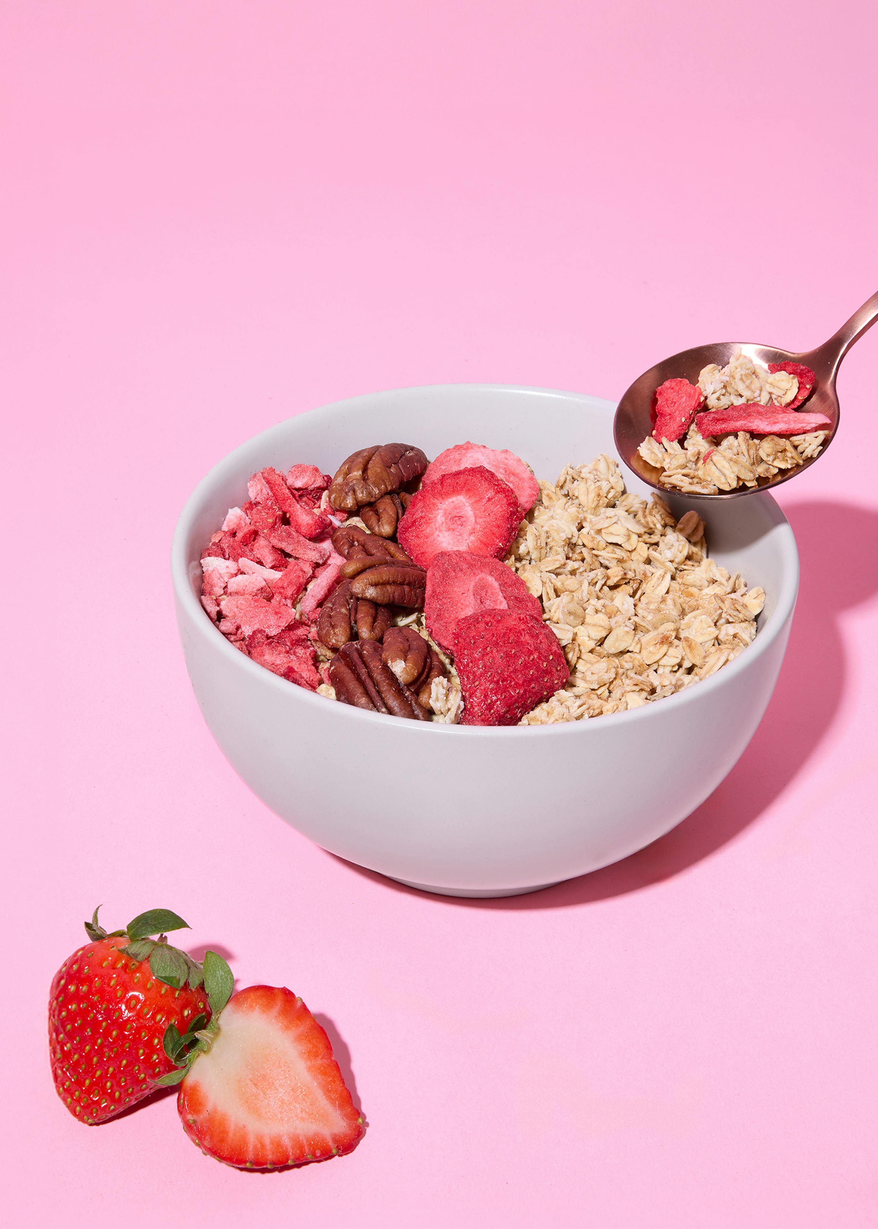 eine weiße schale mit haferflocken, pekannüssen und gefriergetrockneten erdbeeren steht auf einem pinkfarbenen hintergrund, während ein löffel mit haferflocken und erdbeeren über der schale schwebt und frische erdbeeren daneben liegen.