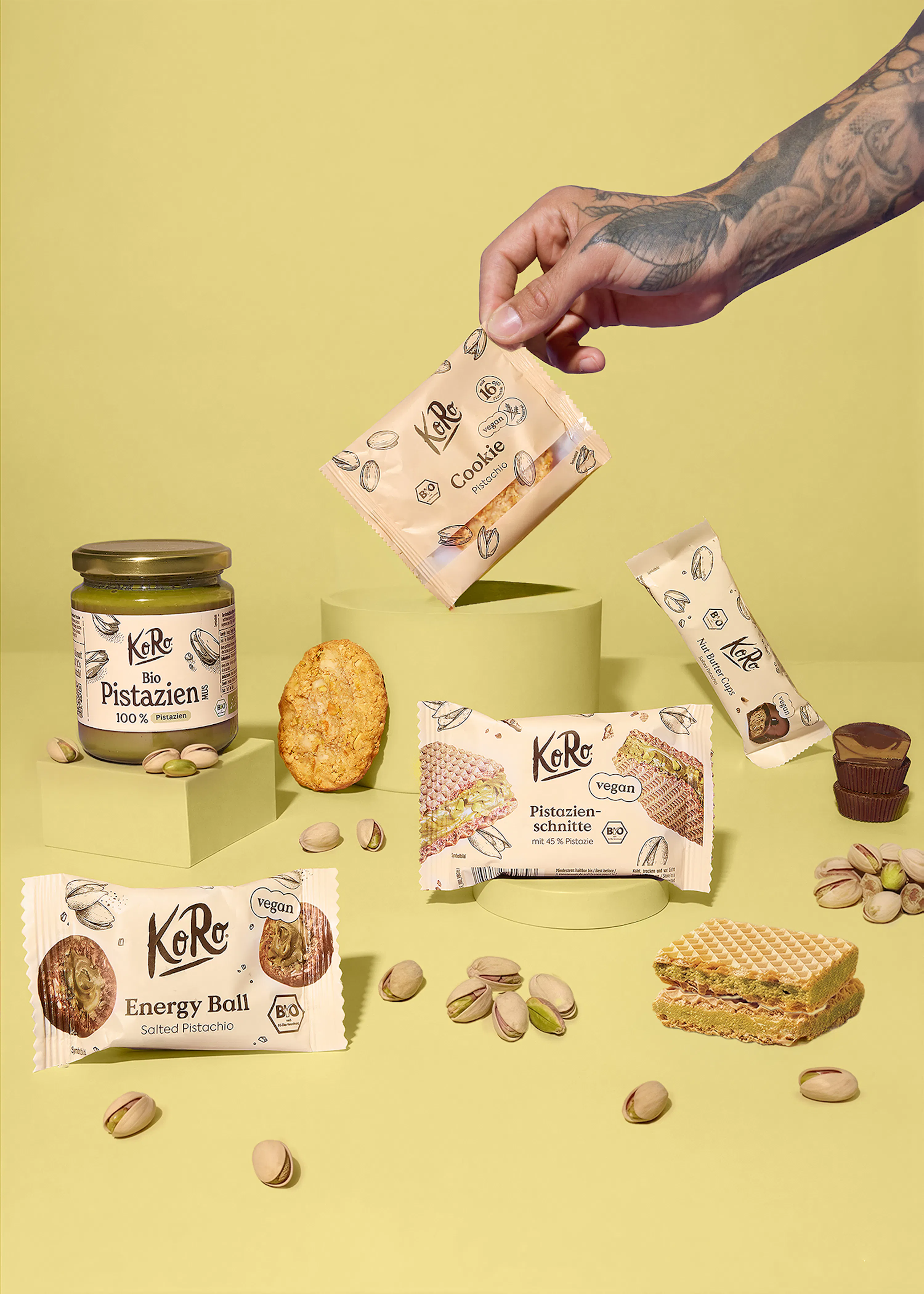 una mano tatuata tiene un pacchetto di biscotti al pistacchio koro, mentre altri prodotti koro al pistacchio, tra cui crema spalmabile, barrette, fette e pistacchi sfusi, sono disposti su uno sfondo giallo.