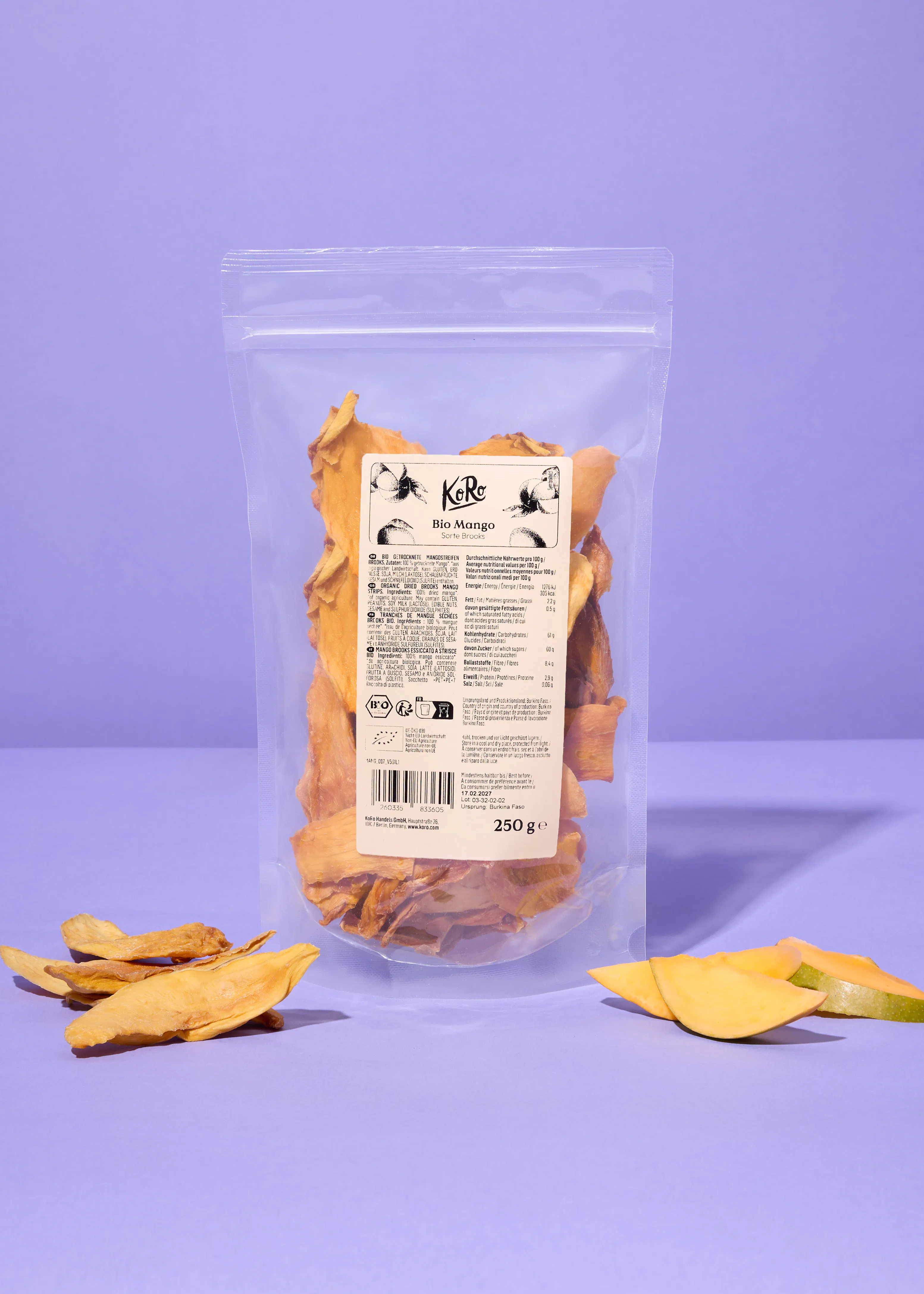 a clear plastic bag of koro bio mango dried mango slices sits on a purple surface, with loose dried mango pieces and fresh mango slices scattered around it.