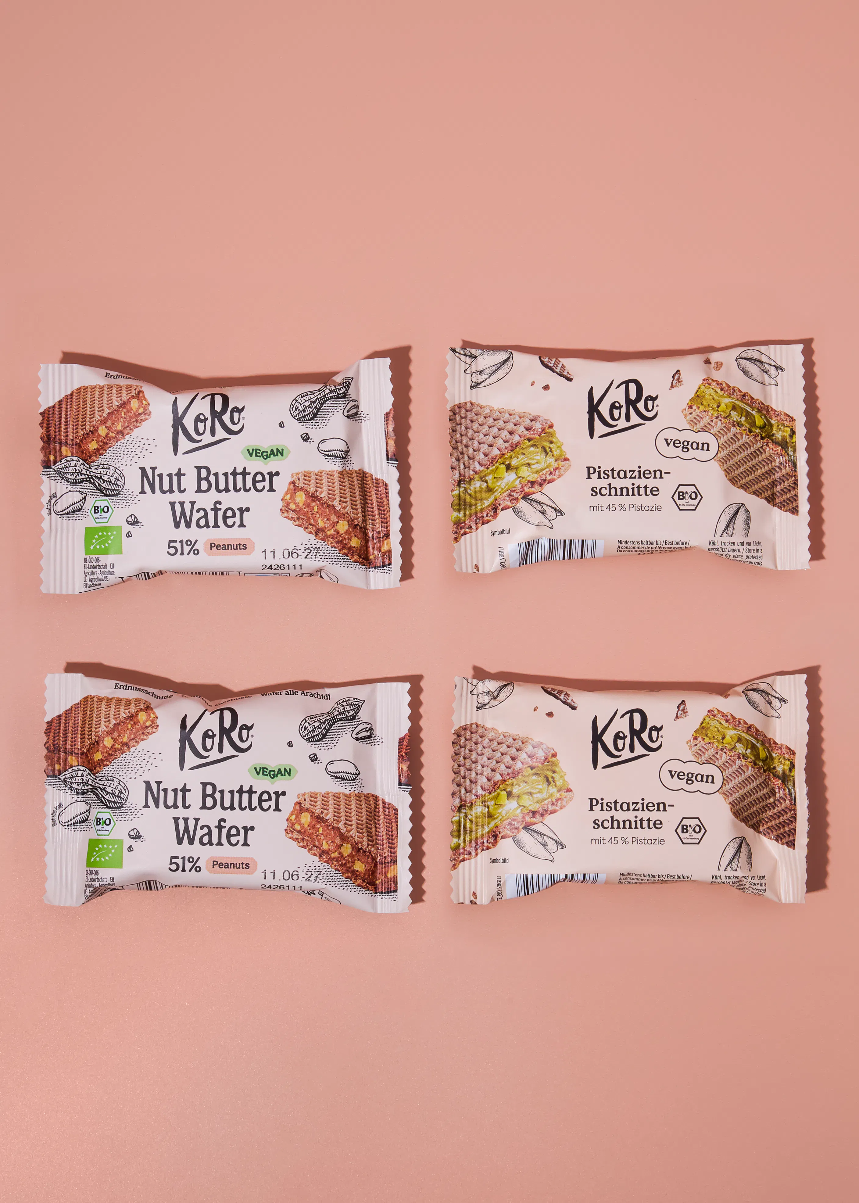 cuatro paquetes de barritas veganas de la marca koro, dos de oblea de mantequilla de cacahuete y dos de oblea de pistacho, están dispuestos en dos filas sobre un fondo rosa claro.