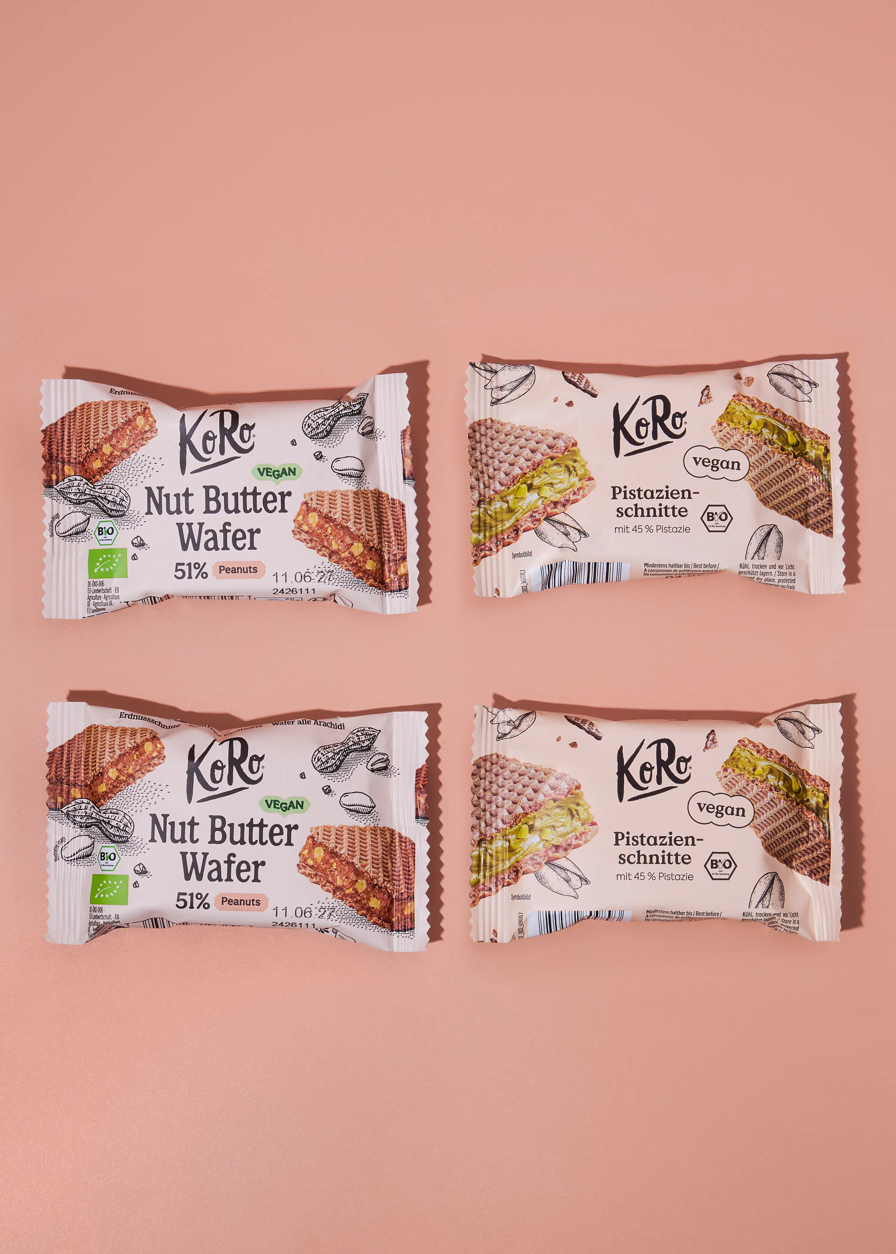 vier verpackte koro vegane waffelsnacks, zwei erdnussbutter-waffeln und zwei pistazien-schnitten, liegen auf einem hellrosa hintergrund.