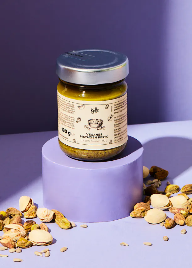 a jar of vegan pistachio pesto sits on a purple podium surrounded by pistachios