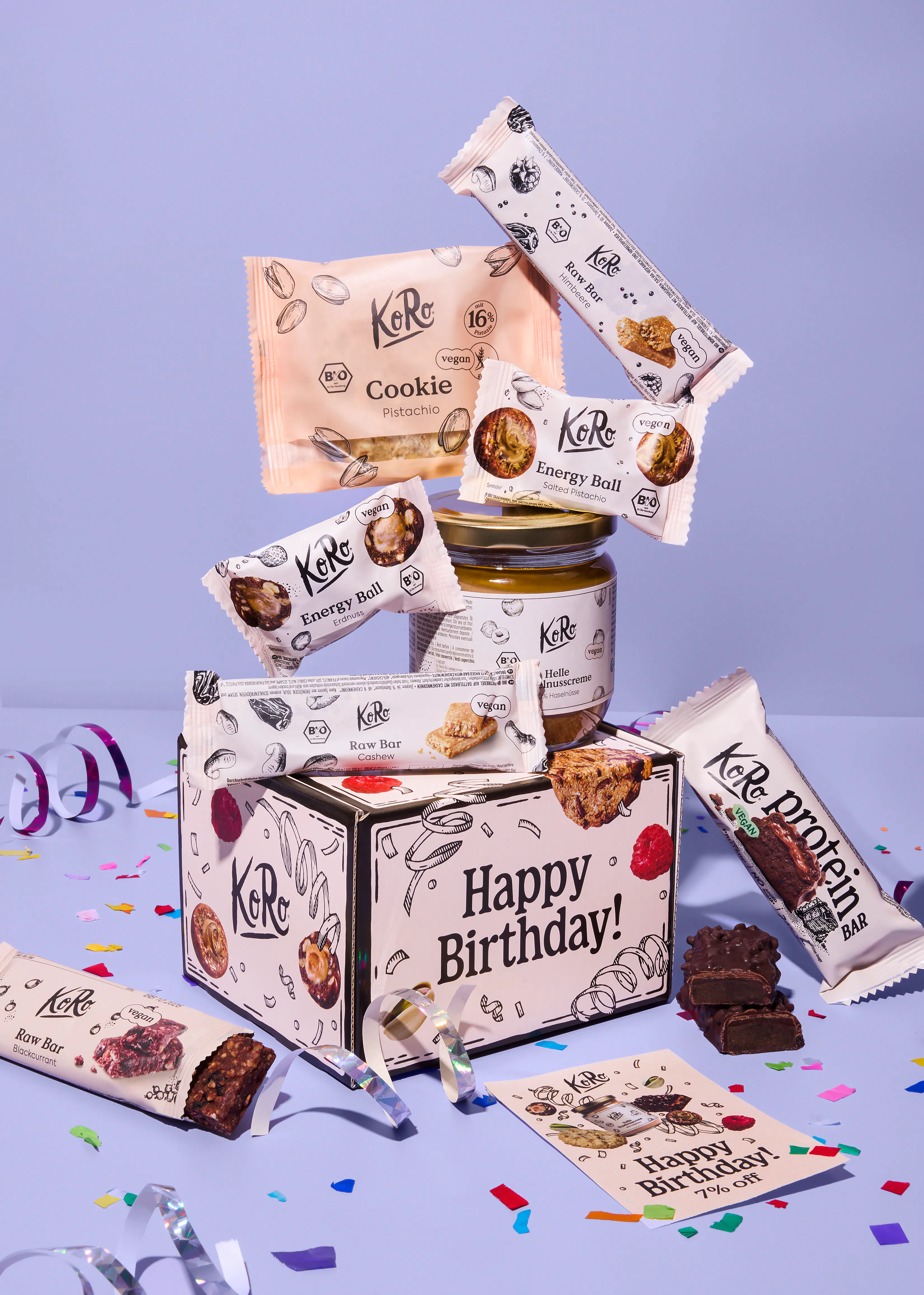 eine koro-geburtstagsbox umgeben von verschiedenen veganen snacks wie riegeln, energy balls und einem glas nusscreme auf einem lila hintergrund mit konfetti.