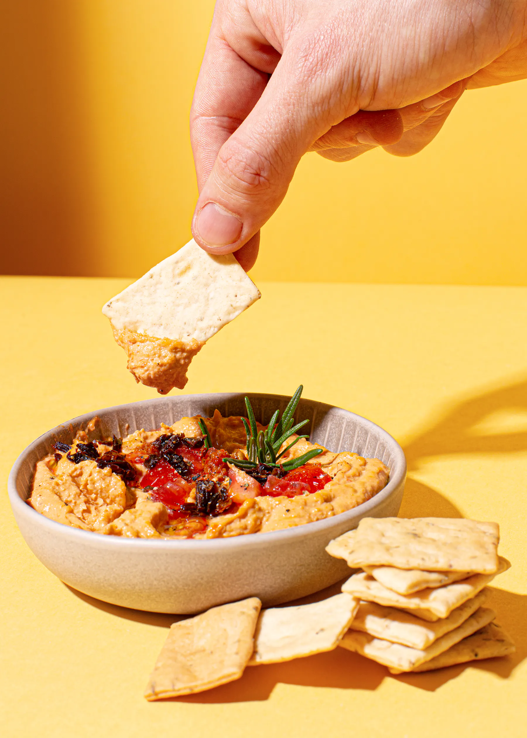 a hand dipping a cracker into a bowl of hummus