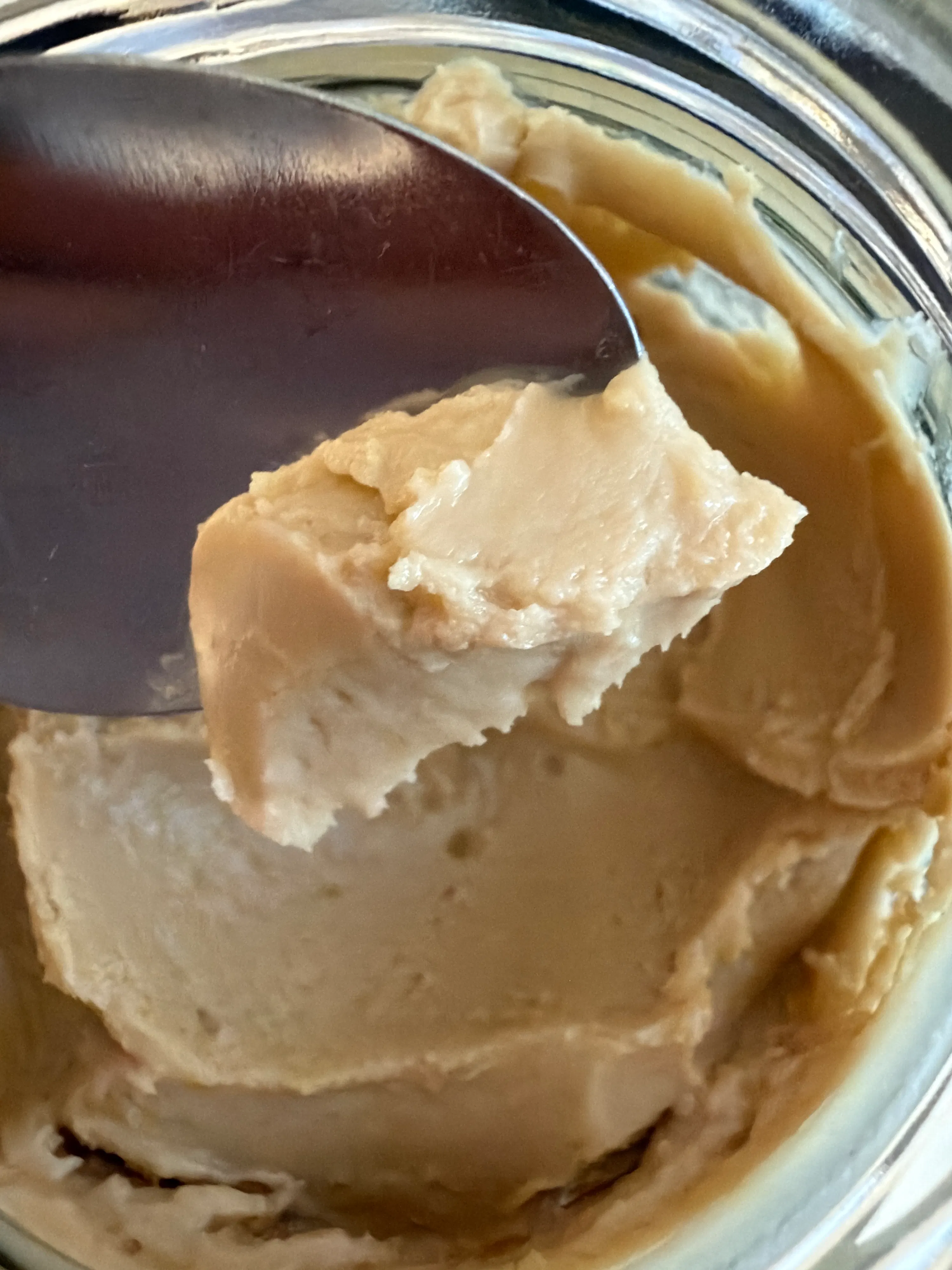 une cuillère en métal prélevant une portion épaisse et crémeuse de couleur beige clair d'un pot.