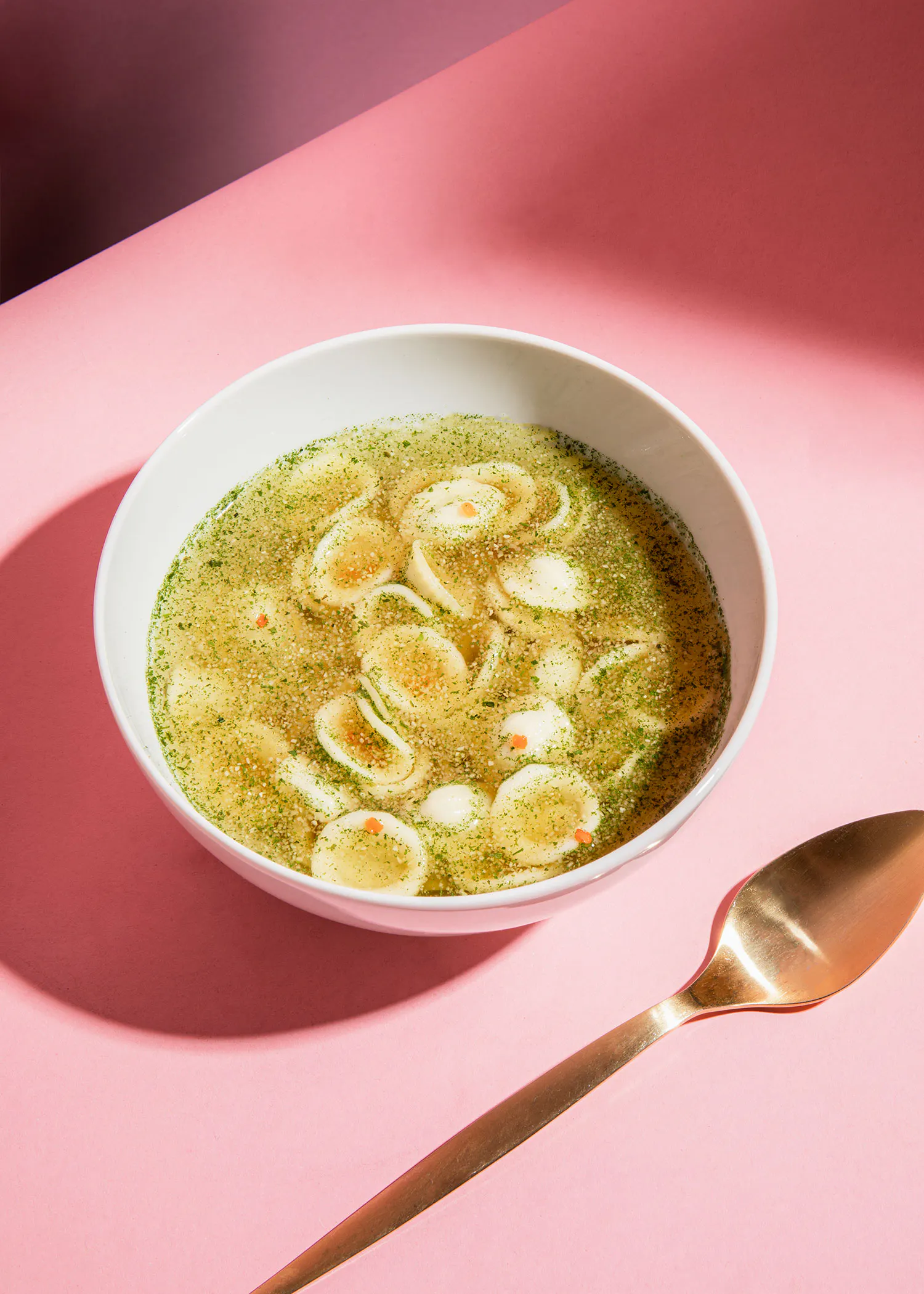 eine weiße Schüssel Suppe mit einem goldenen Löffel auf einem rosa Tisch