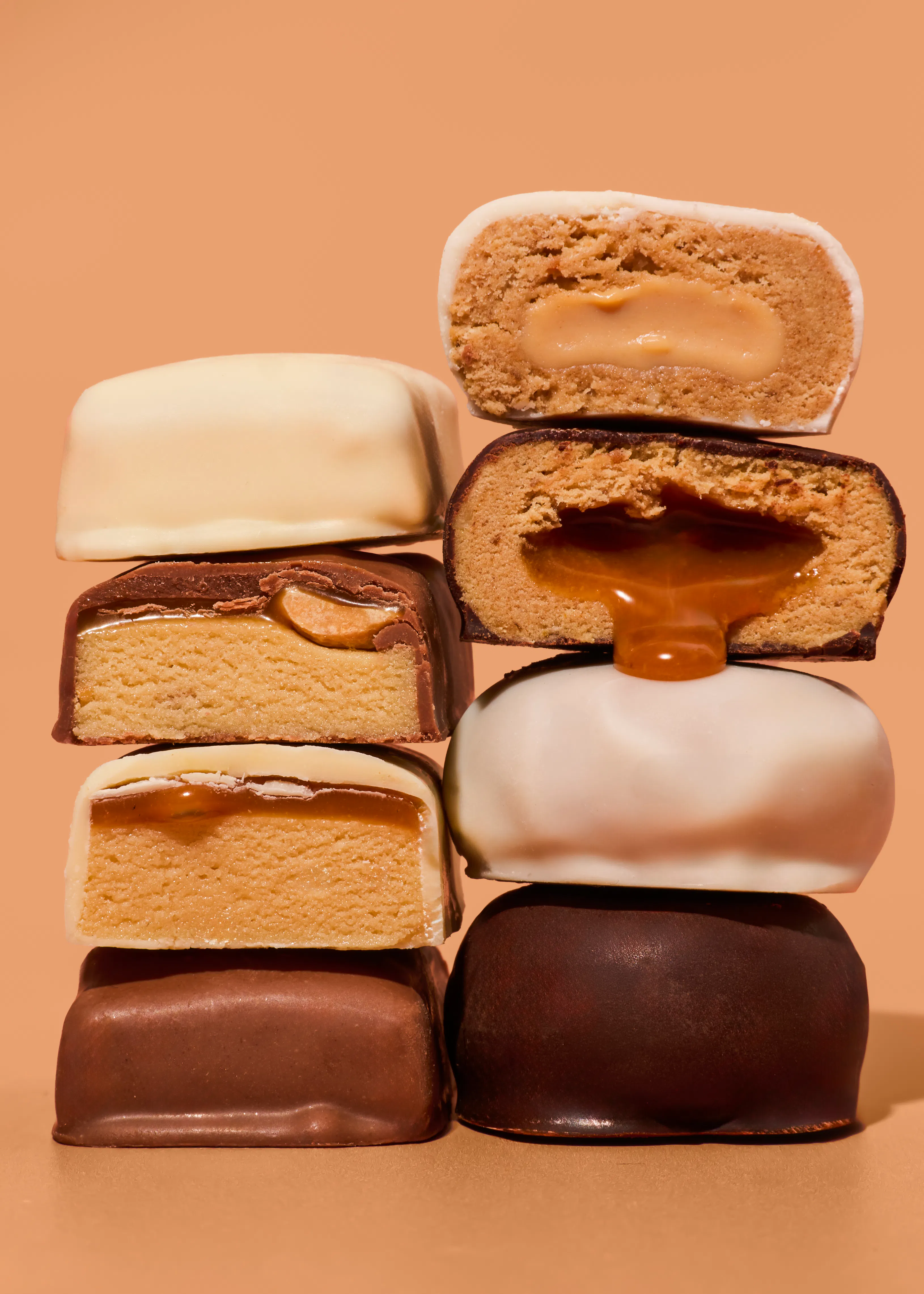 una pila di barrette proteiche tagliate a metà che mostrano diversi ripieni cremosi e al caramello ricoperte di cioccolato bianco o fondente su sfondo color sabbia.
