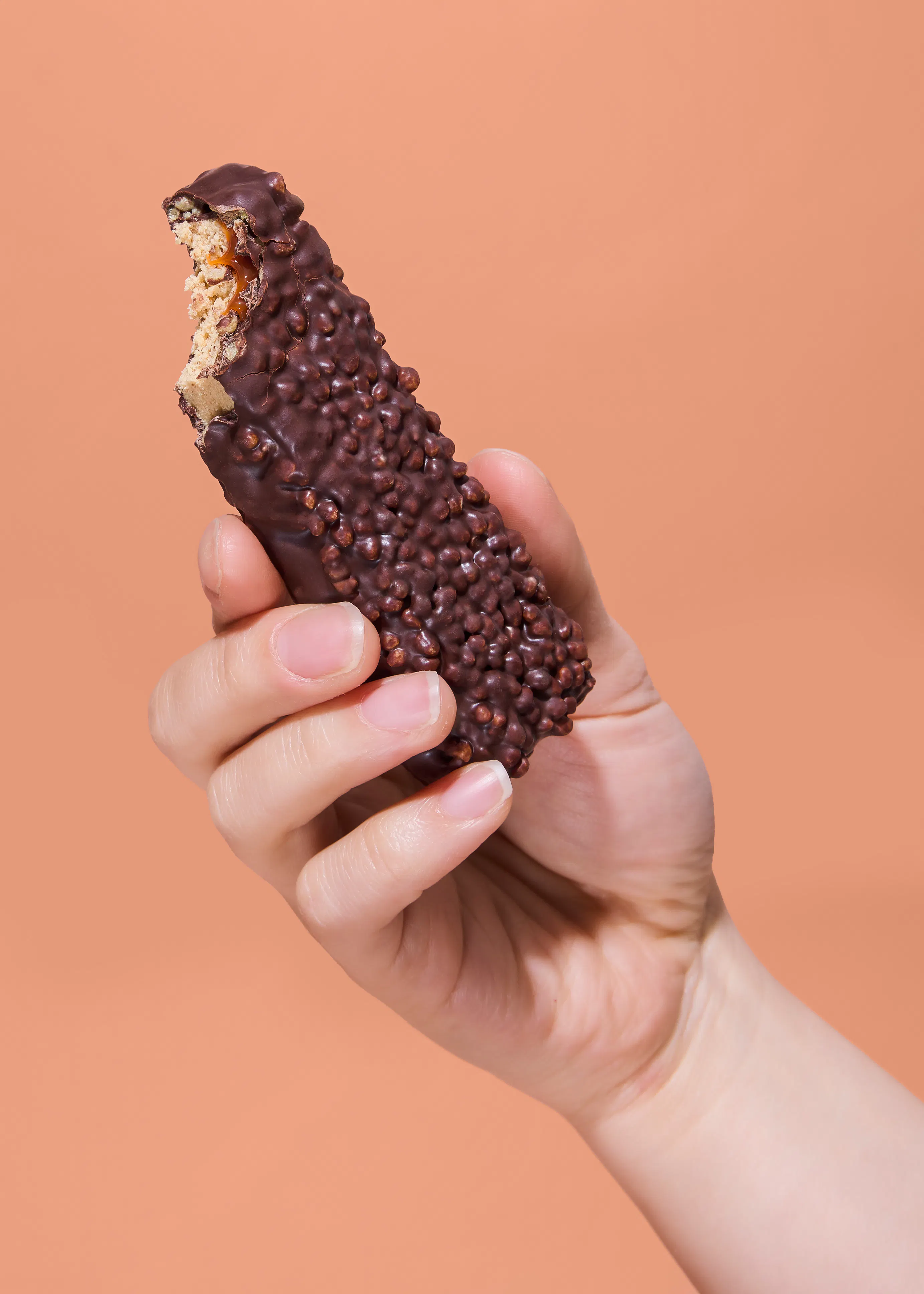 una mano tiene una barretta di cioccolato con un morso che rivela un ripieno croccante e caramellato, su sfondo marrone chiaro.