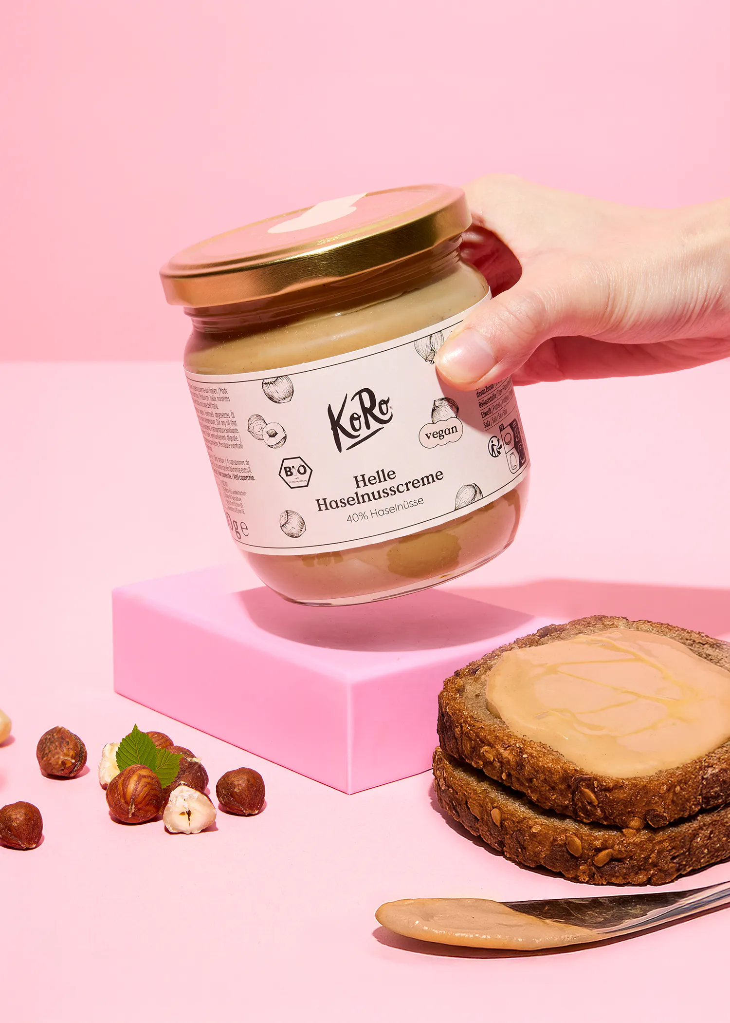 eine hand hält ein glas helle haselnusscreme von koro über einem brot mit aufstrich, umgeben von haselnüssen und einem messer auf einer rosa oberfläche.