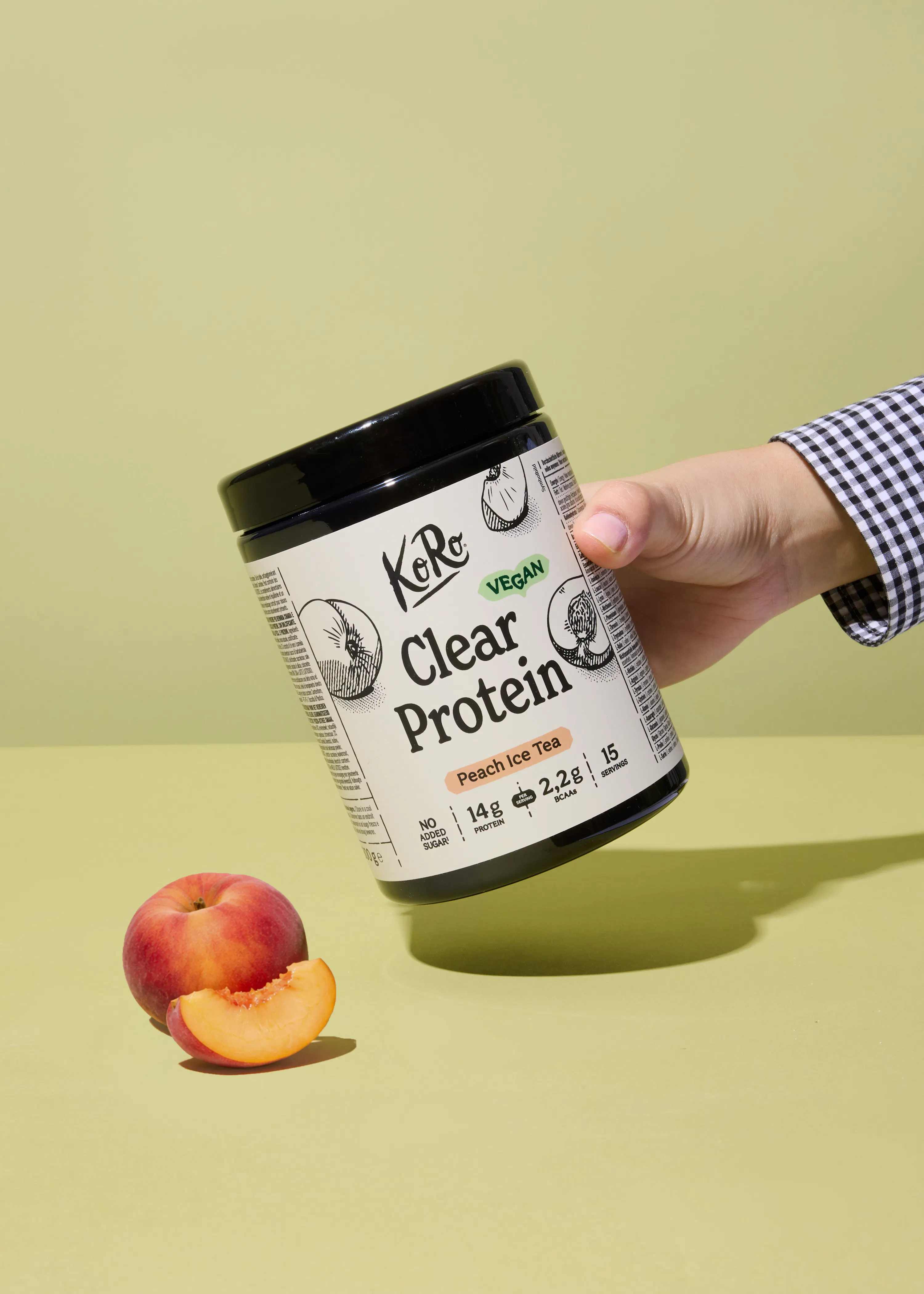 a hand in a checkered shirt holds a koro clear protein peach ice tea container, labeled vegan with 14g protein, next to a whole peach and a peach slice on a light green background.
