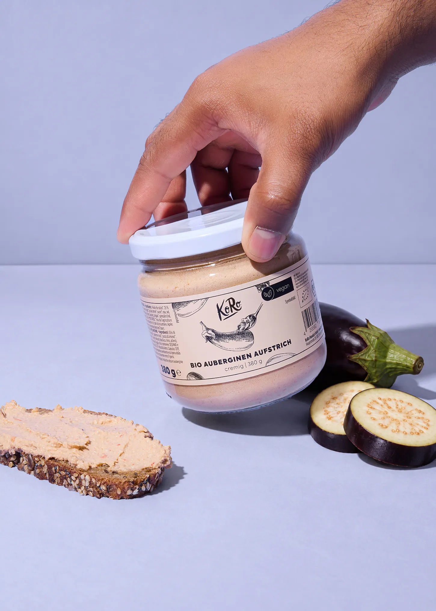 eine hand hält ein glas koro bio-auberginen-aufstrich über eine scheibe brot mit aufstrich und frischen auberginenstücken.