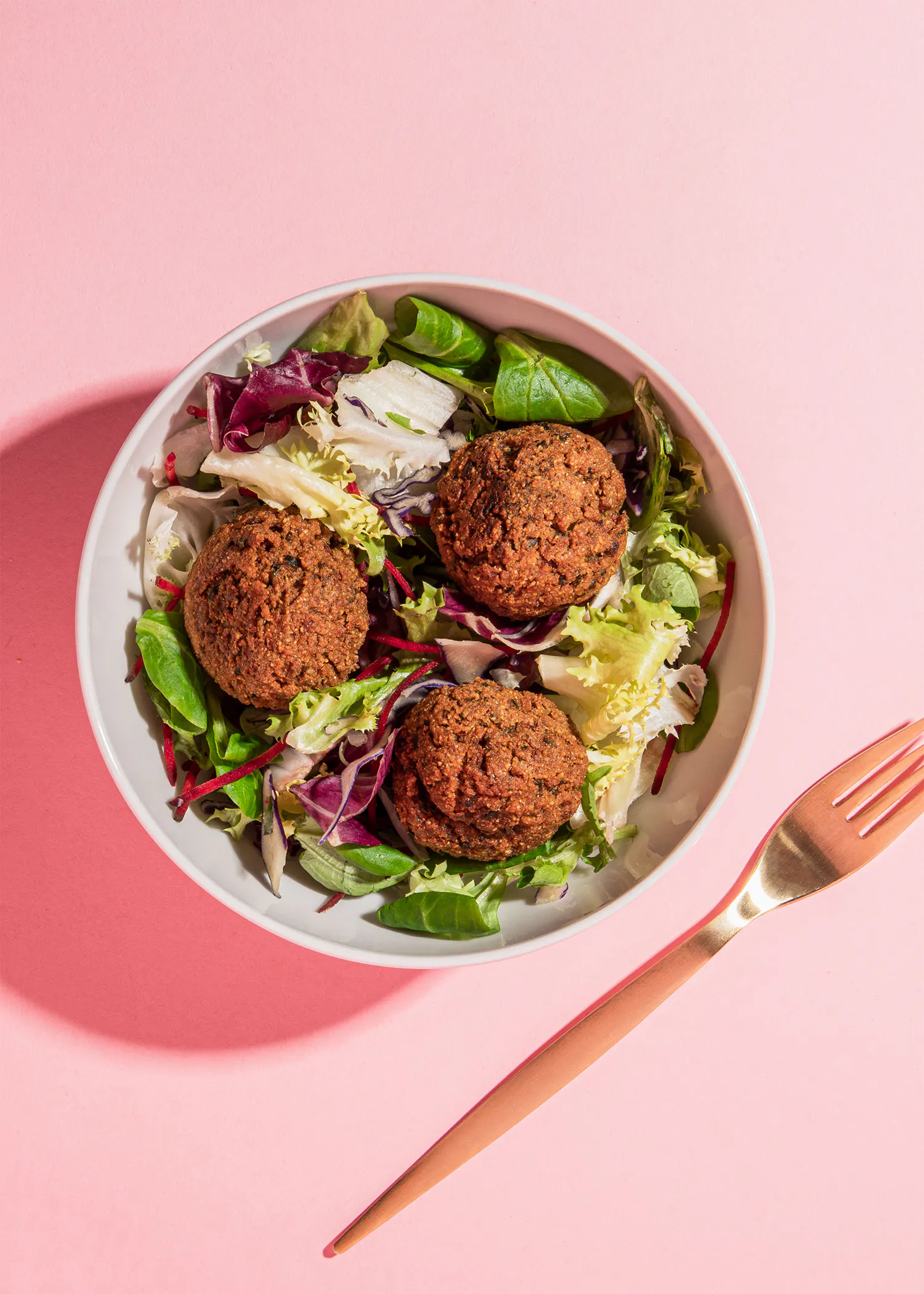 a bowl of falafel salad with a fork on a pink background