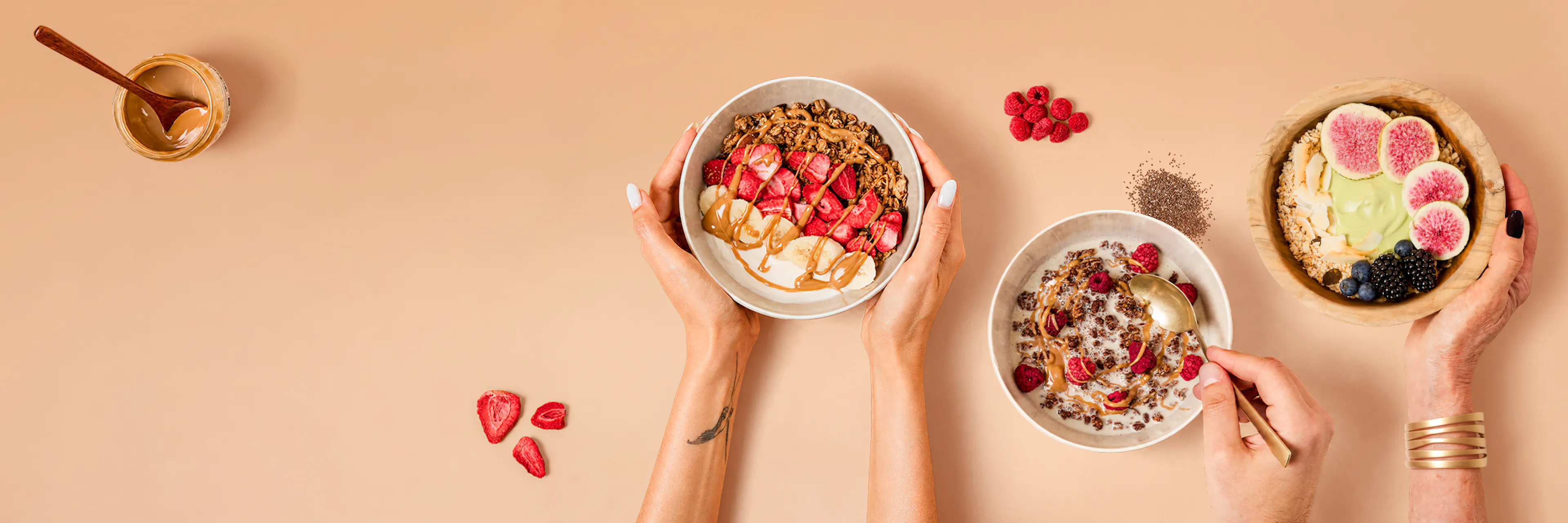drei frühstücksbowls mit müsli, früchten und nussbutter werden auf einem beigen hintergrund präsentiert, wobei hände zwei der schüsseln halten und eine person eine davon isst.