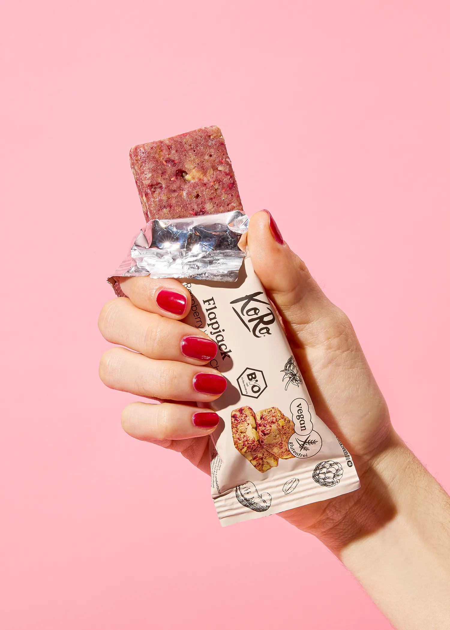 une main avec des ongles rouges tient une barre de céréales tropicale