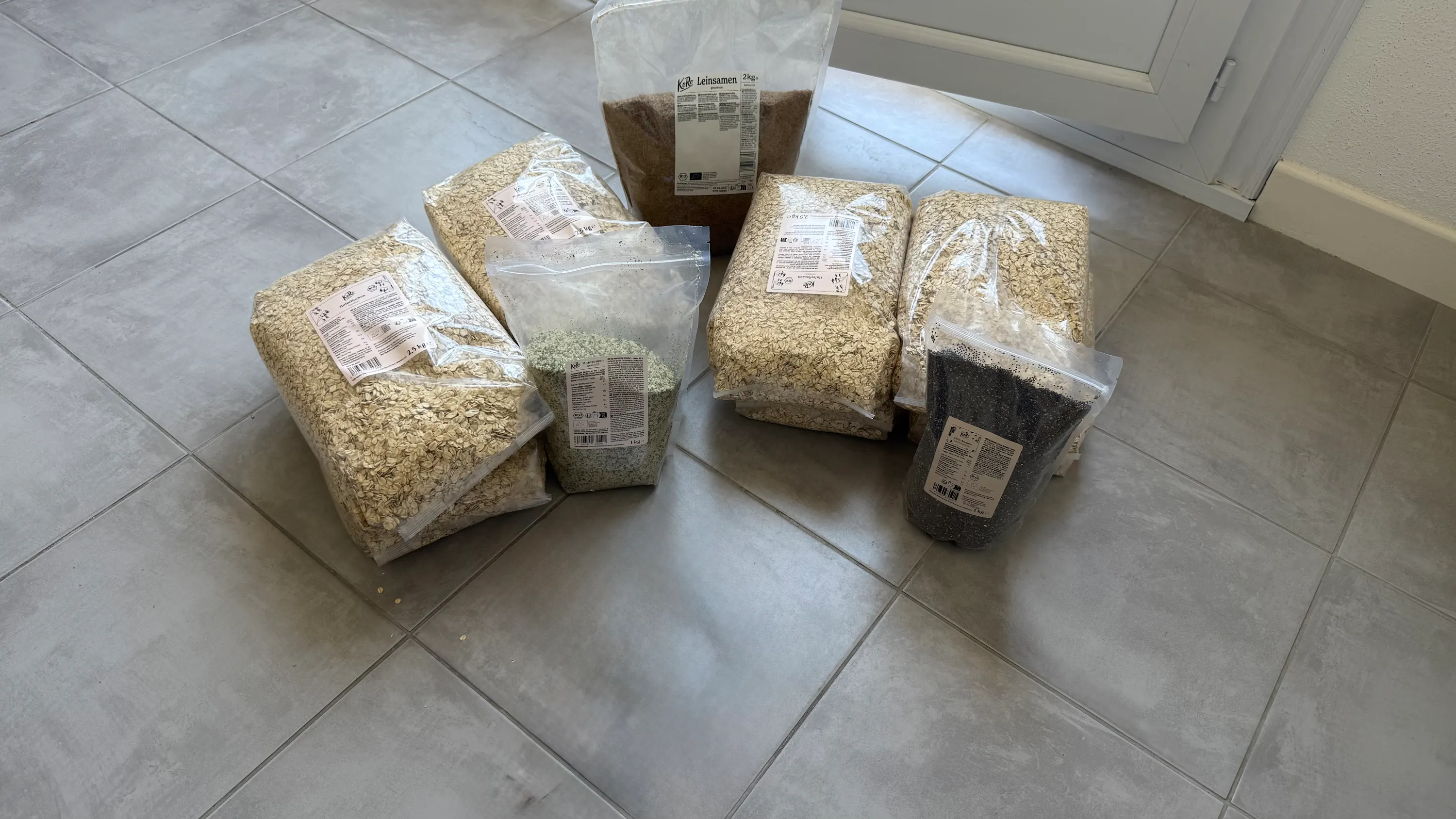diversi sacchetti di plastica trasparenti pieni di fiocchi d'avena, semi di lino e semi di chia sono disposti su un pavimento piastrellato grigio.