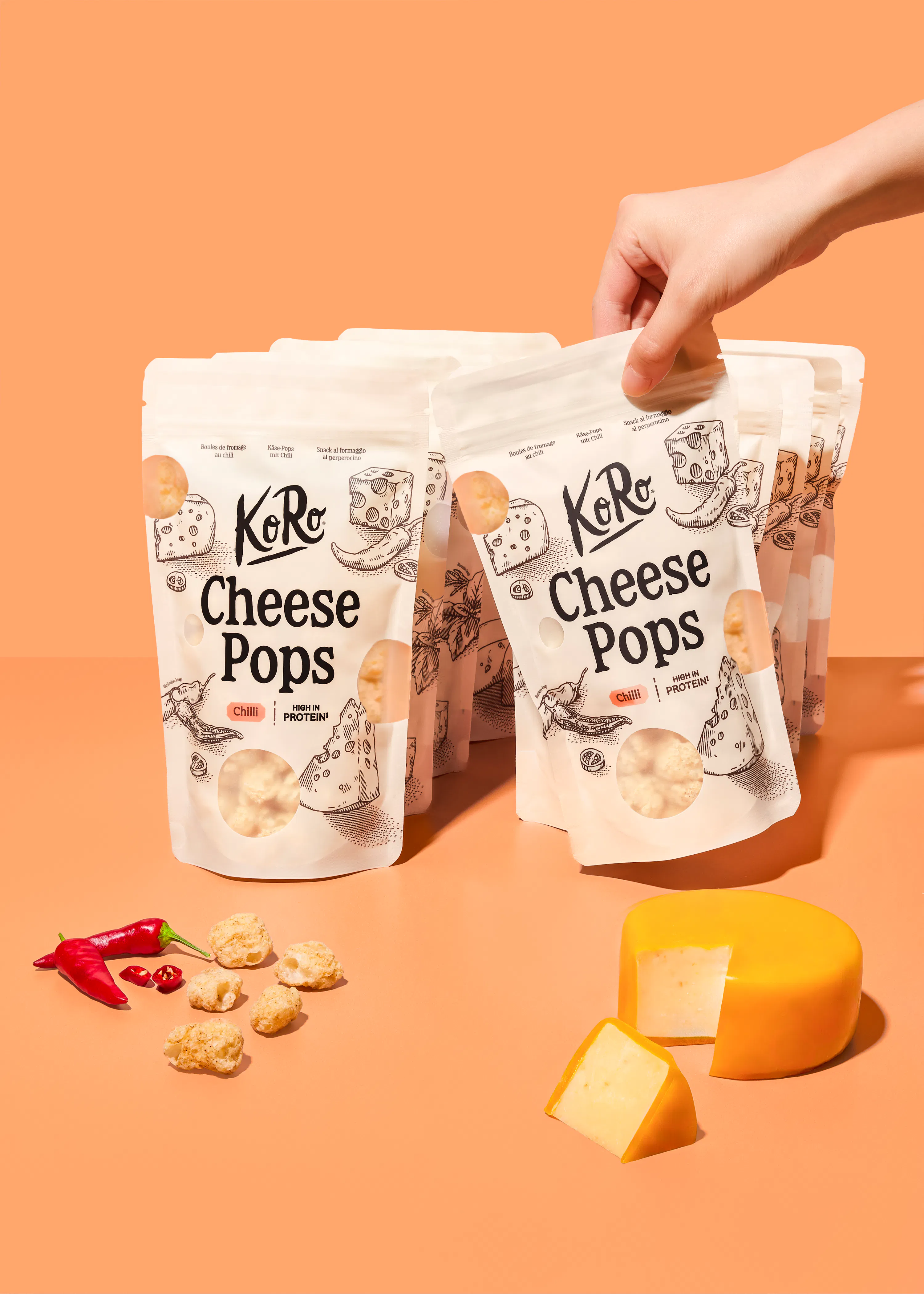 una mano solleva una confezione di koro cheese pops al peperoncino su uno sfondo color pesca, con accanto una forma di formaggio e peperoncini freschi.