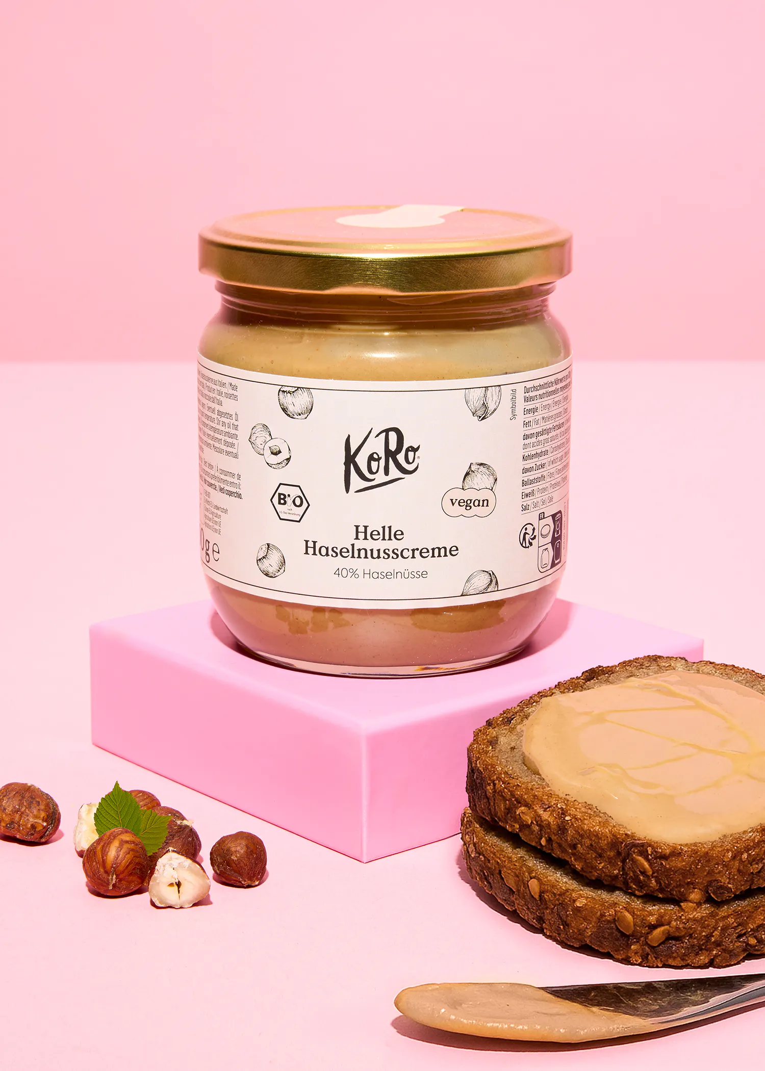 ein glas heller haselnusscreme von koro auf einem rosa block, daneben ganze und geschälte haselnüsse sowie brotscheiben mit dem aufstrich und ein messer.