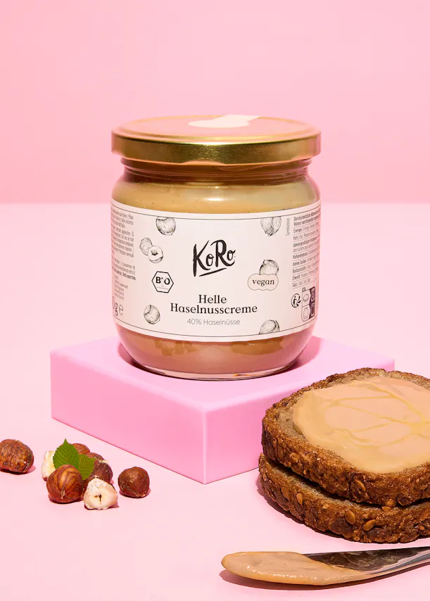 ein glas heller haselnusscreme von koro auf einem rosa block, daneben ganze und geschälte haselnüsse sowie brotscheiben mit dem aufstrich und ein messer.