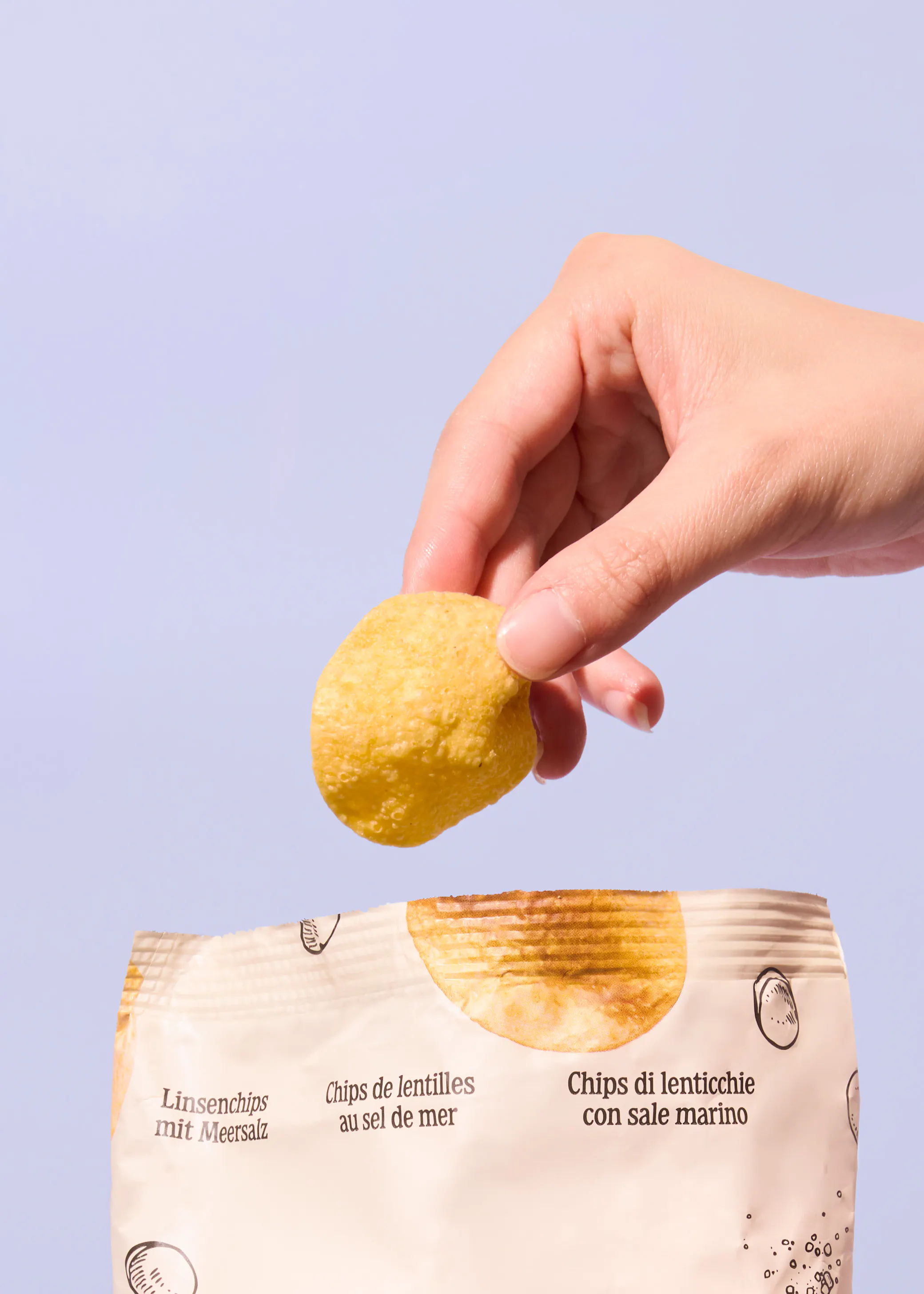 eine hand hält einen einzelnen linsenchip über einer chipstüte vor einem hellvioletten hintergrund.