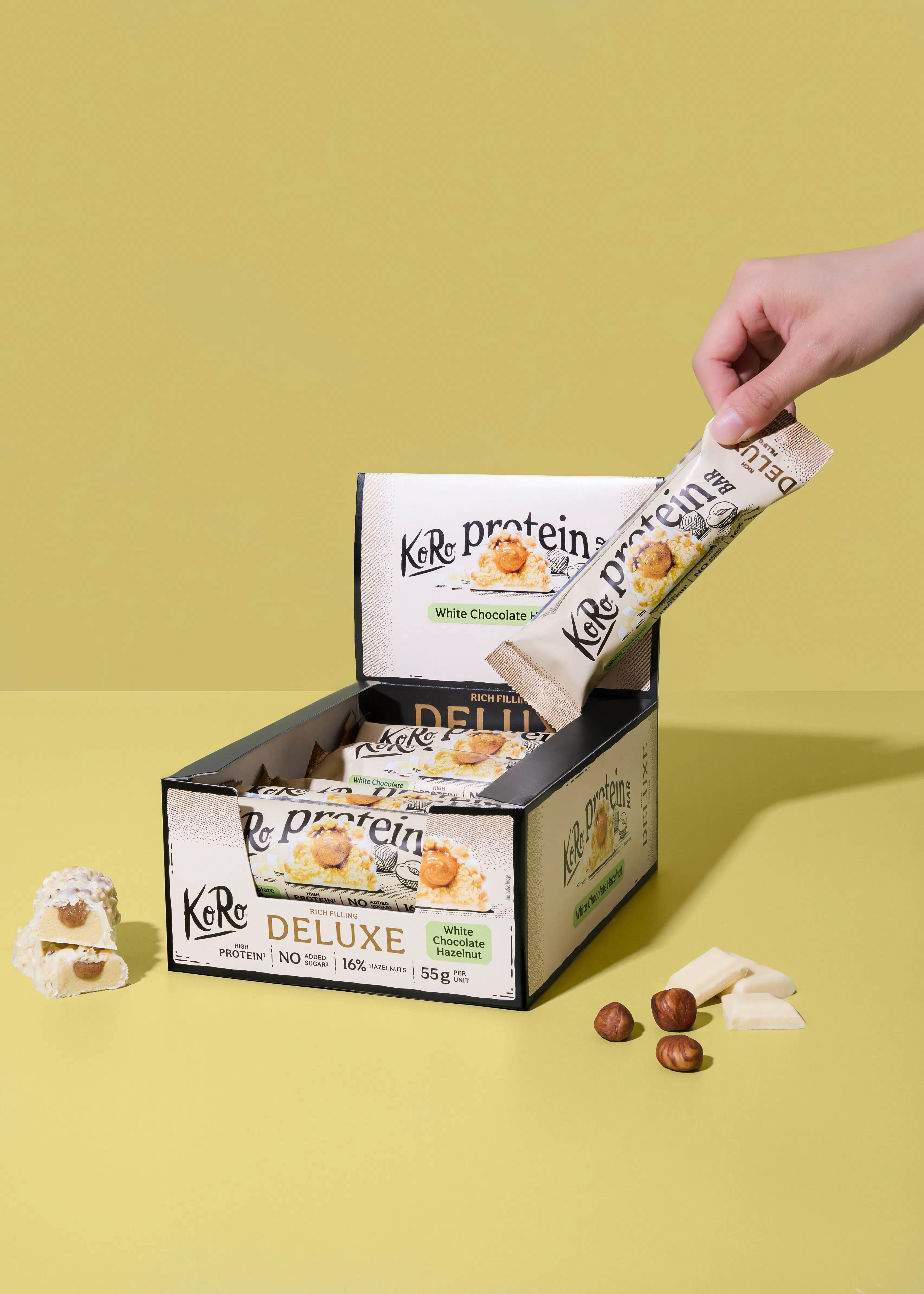 une main prend une barre protéinée koro deluxe au chocolat blanc et noisette d'une boîte, avec des noisettes, du chocolat blanc et une barre coupée à côté.