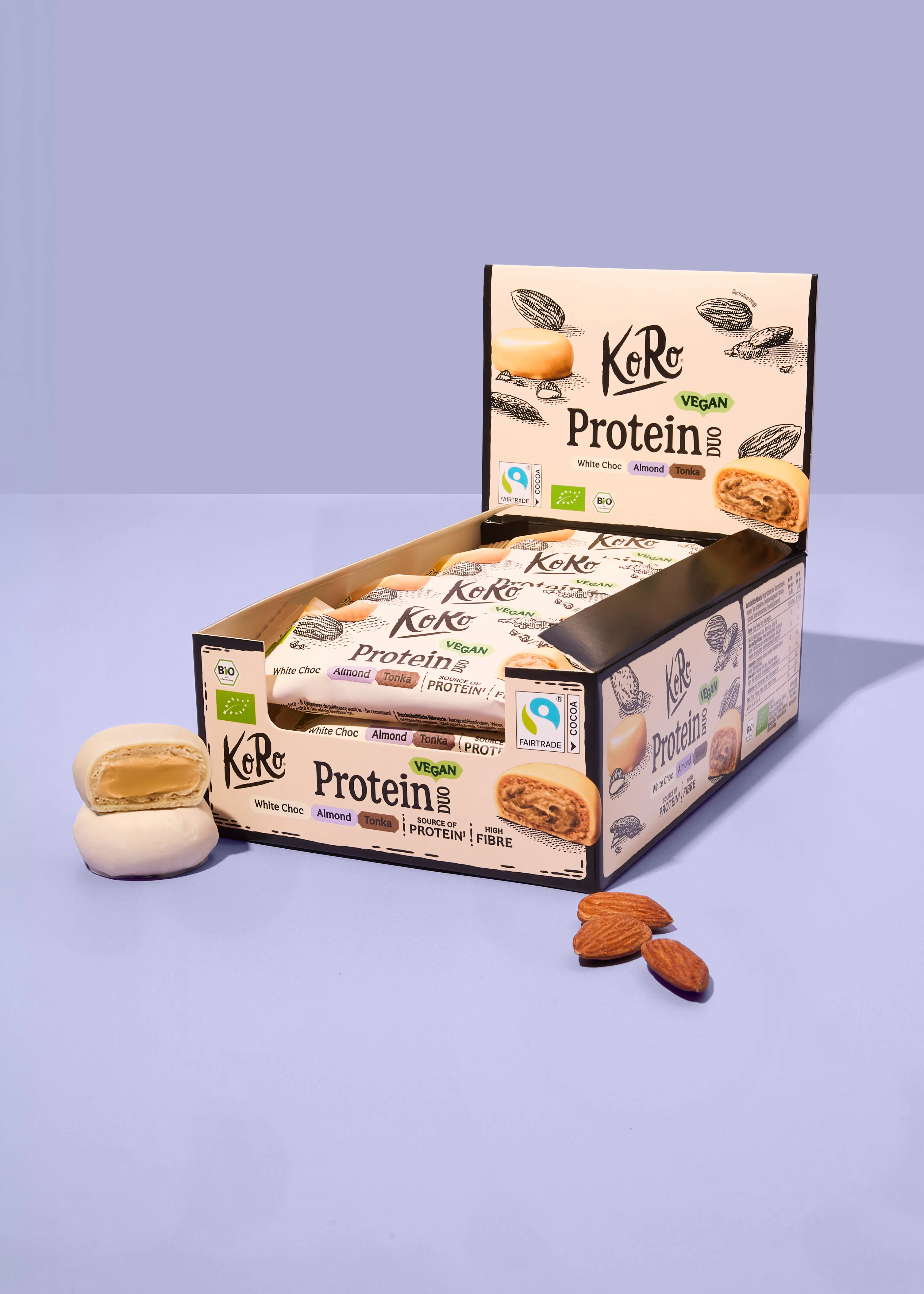 eine geöffnete verkaufsverpackung mit koro vegan protein duo riegeln in der geschmacksrichtung white choc almond tonka, daneben liegen zwei halbierte riegel und drei mandeln auf einem hellvioletten hintergrund.